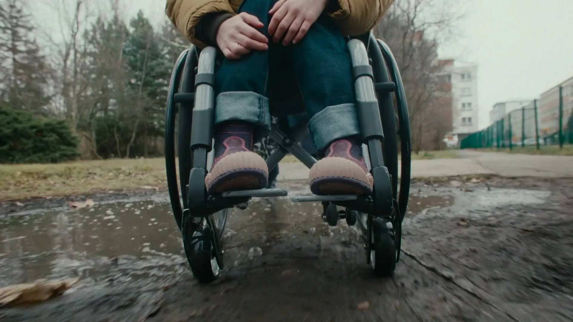 A child in a wheelchair is pushed through a muddy puddle on a path in a park on an overcast day.