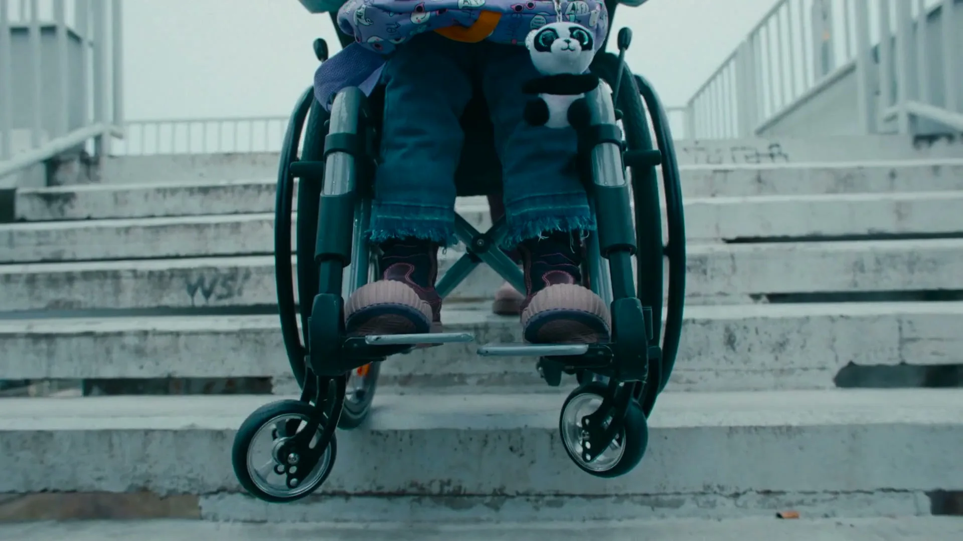 A child in a wheelchair, wearing blue jeans and pink shoes, is seen from the waist down, attempting to ascend a concrete staircase outdoors.