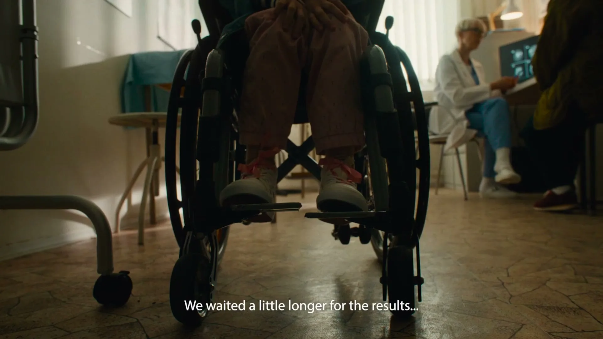 A low-angle shot shows a child's legs in a wheelchair, with a doctor working at a computer in the background of a hospital room.