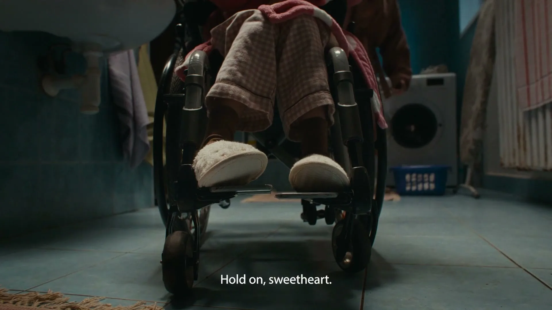 A low-angle shot shows the feet of a person in a wheelchair, wearing checkered pajamas and fuzzy slippers, as another person walks past in a bathroom.