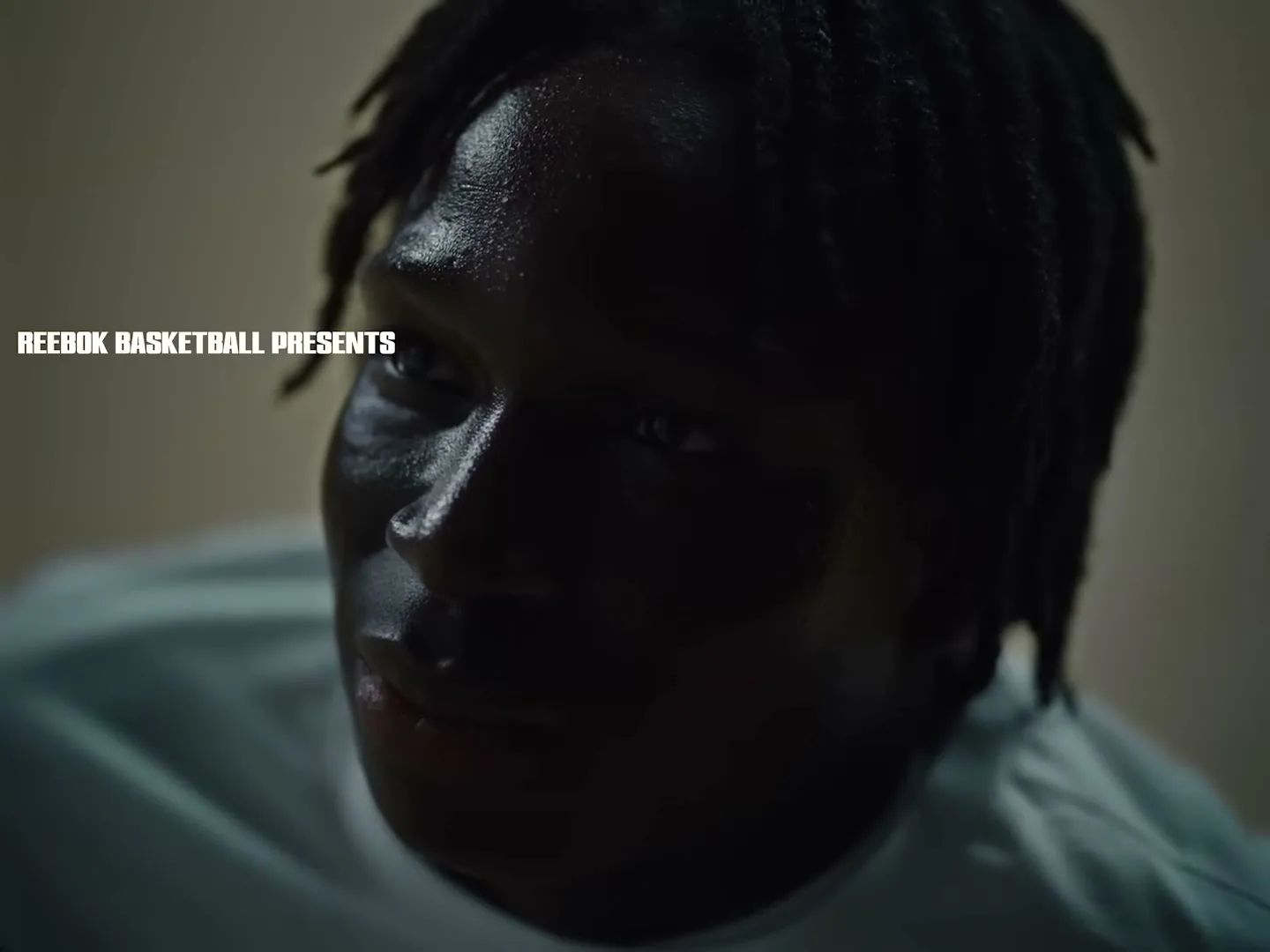 A young Black man with dreadlocks, wearing a light blue shirt, looks intently to the side in a dimly lit room.