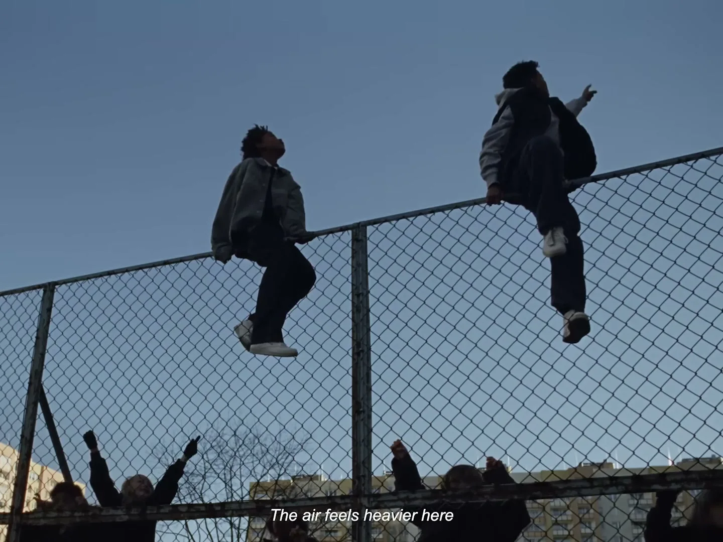 Two young men sit atop a chain-link fence, silhouetted against a clear blue sky, while others stand below, looking up.