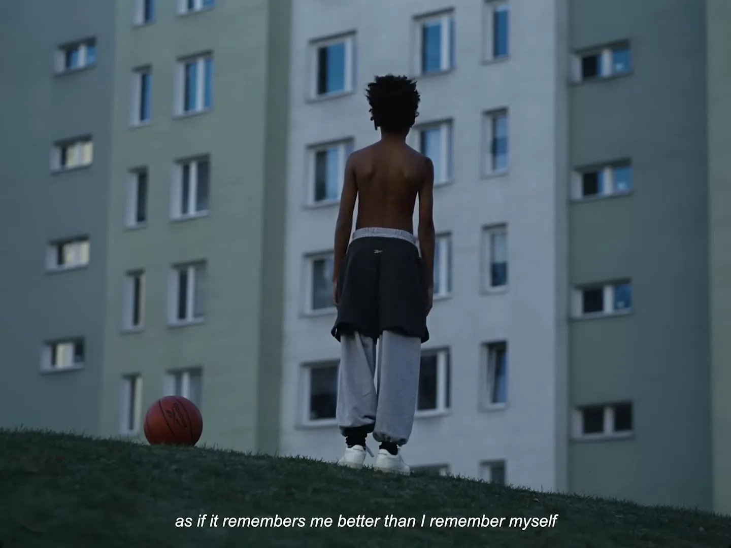 A young boy stands shirtless on a grassy hill, looking towards a tall apartment building, with a basketball resting beside him.