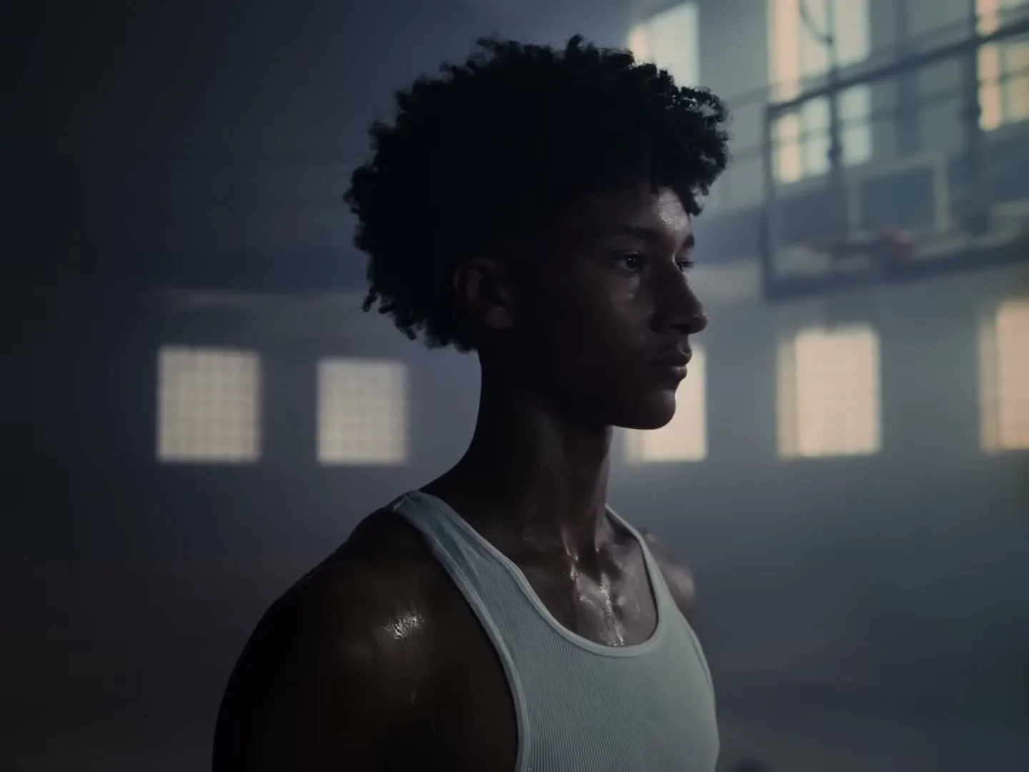 A sweaty male basketball player in a white tank top stands in a dimly lit gym, turning his head to face the camera.