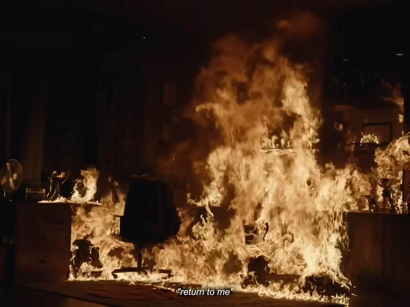 An office chair sits in the middle of a dark office, surrounded by large, bright flames that engulf the room.