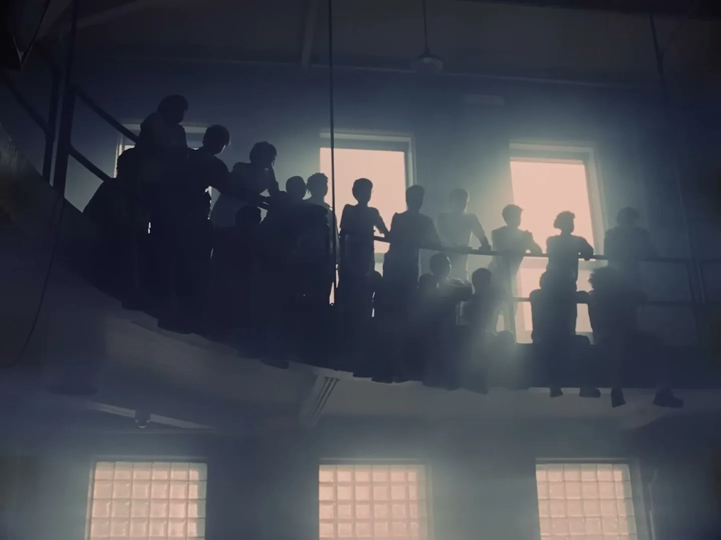 A group of people are silhouetted on a curved balcony, looking out towards bright windows in a large, dark interior space.