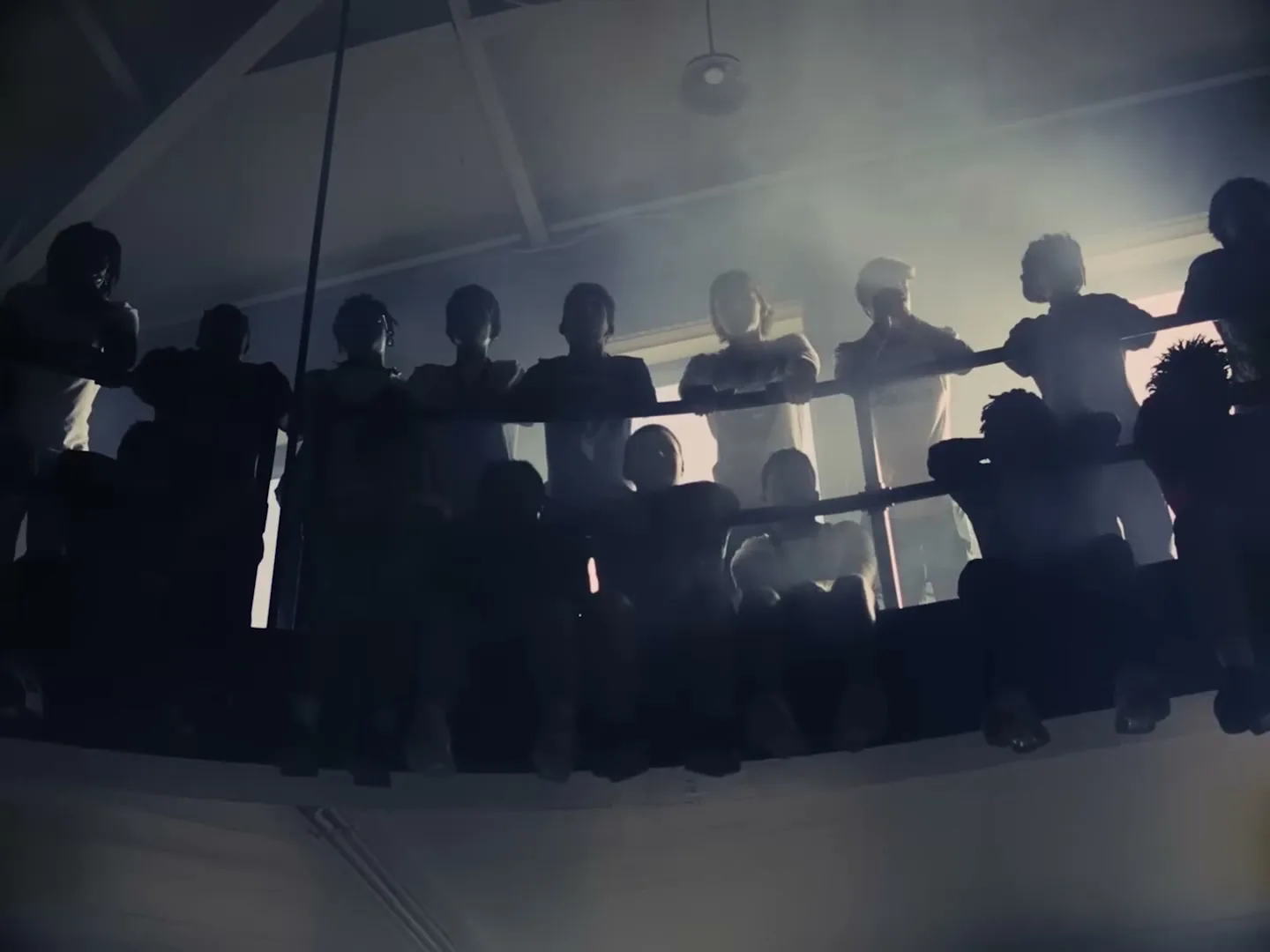 A group of people, silhouetted by bright backlighting, stand around a boxing ring in a dark, smoky gym.