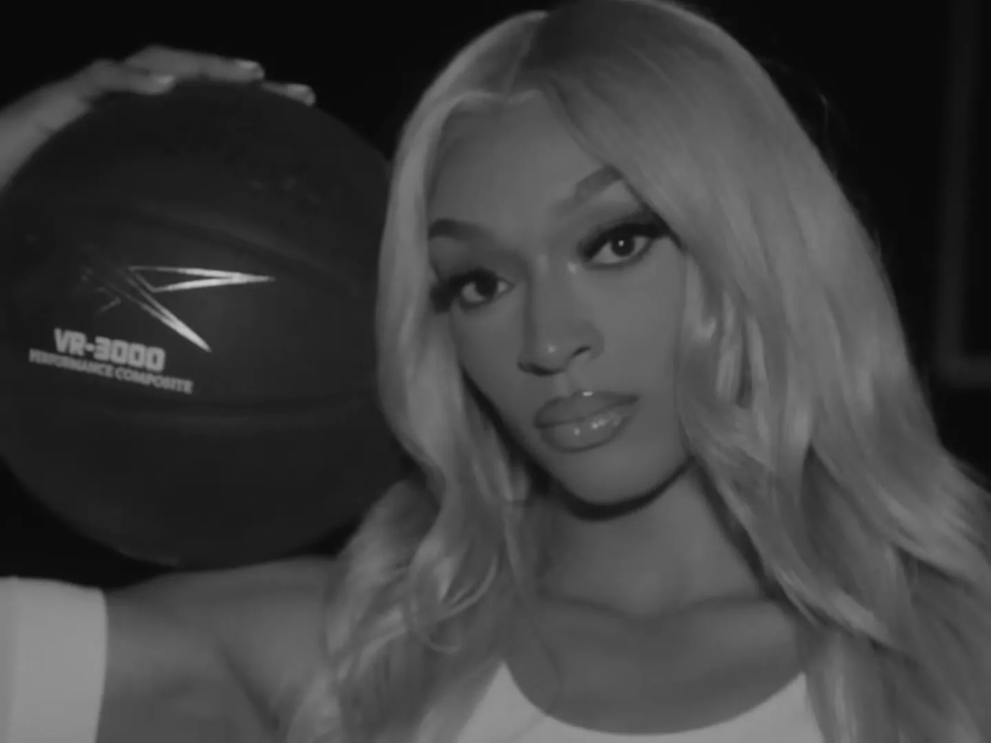 A young woman with long blonde hair holds a basketball over her shoulder, looking directly at the camera in a black and white shot.