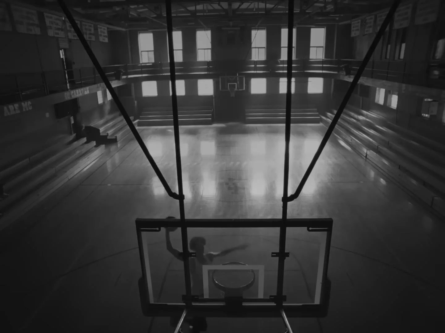 A lone basketball player dunks a ball into a hoop in a dimly lit, empty gymnasium, shot in black and white.