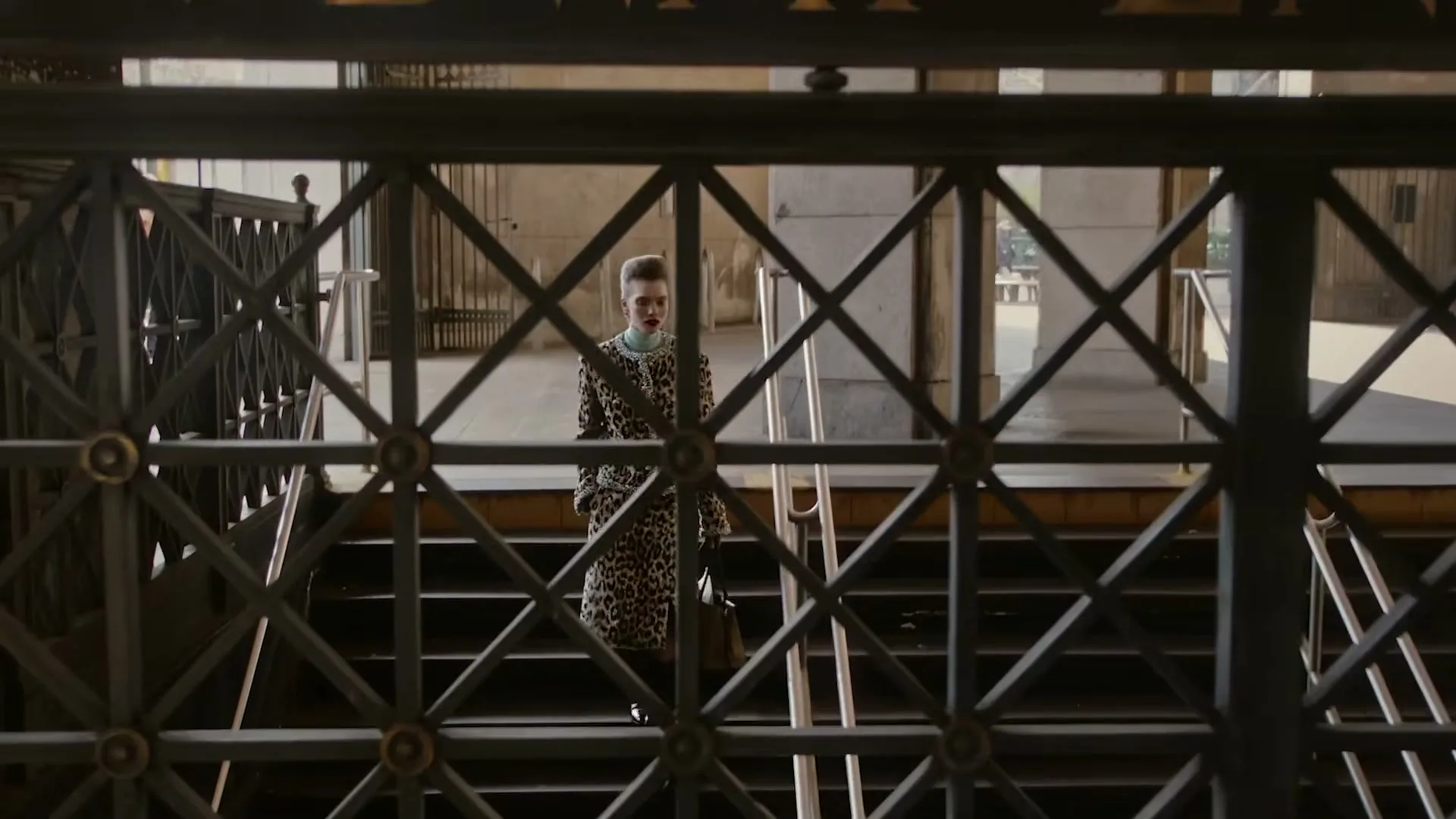 A woman in a leopard print coat stands at the bottom of a subway entrance, looking up through a metal gate.