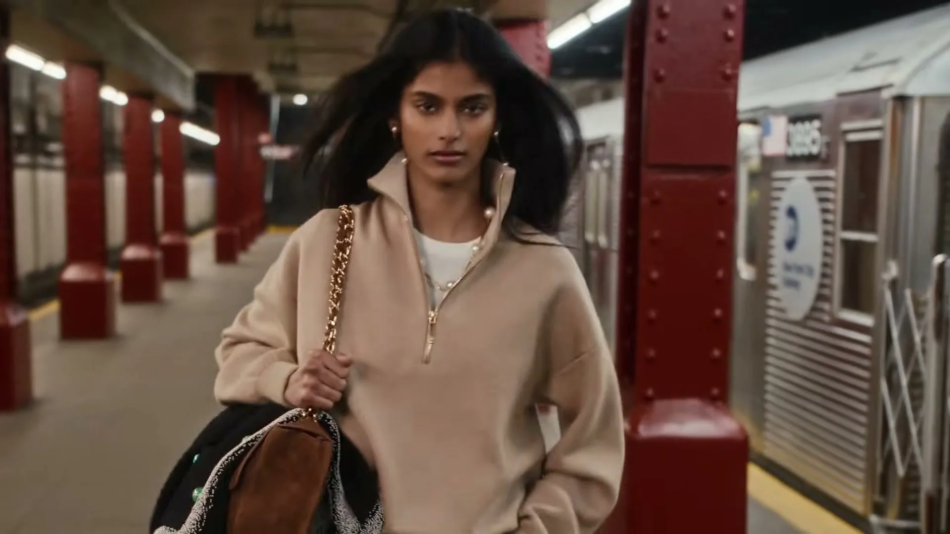 A young woman with dark hair and a beige top walks towards the camera on a subway platform, carrying a bag.