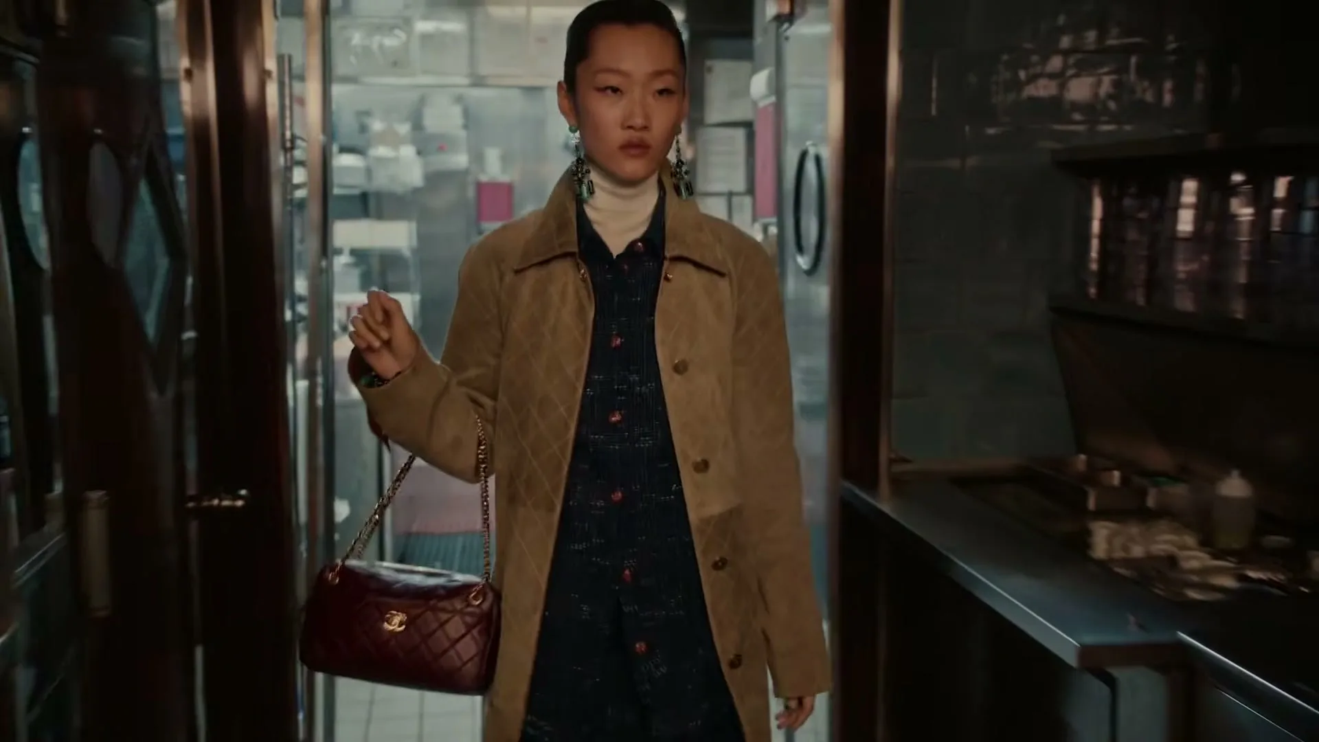 A young Asian woman in a quilted coat and carrying a red handbag walks through a restaurant kitchen.