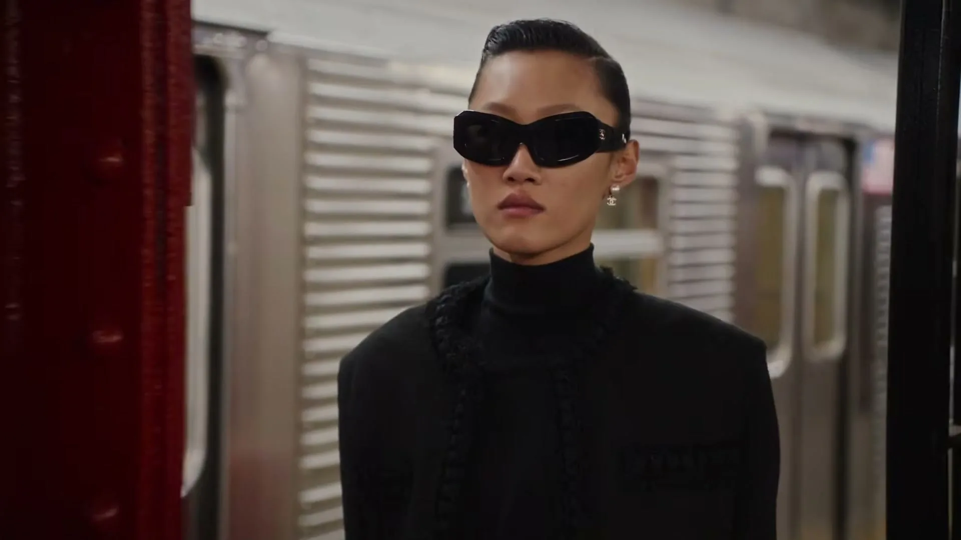 A female model in black clothing and sunglasses stands on a subway platform as a train passes by.