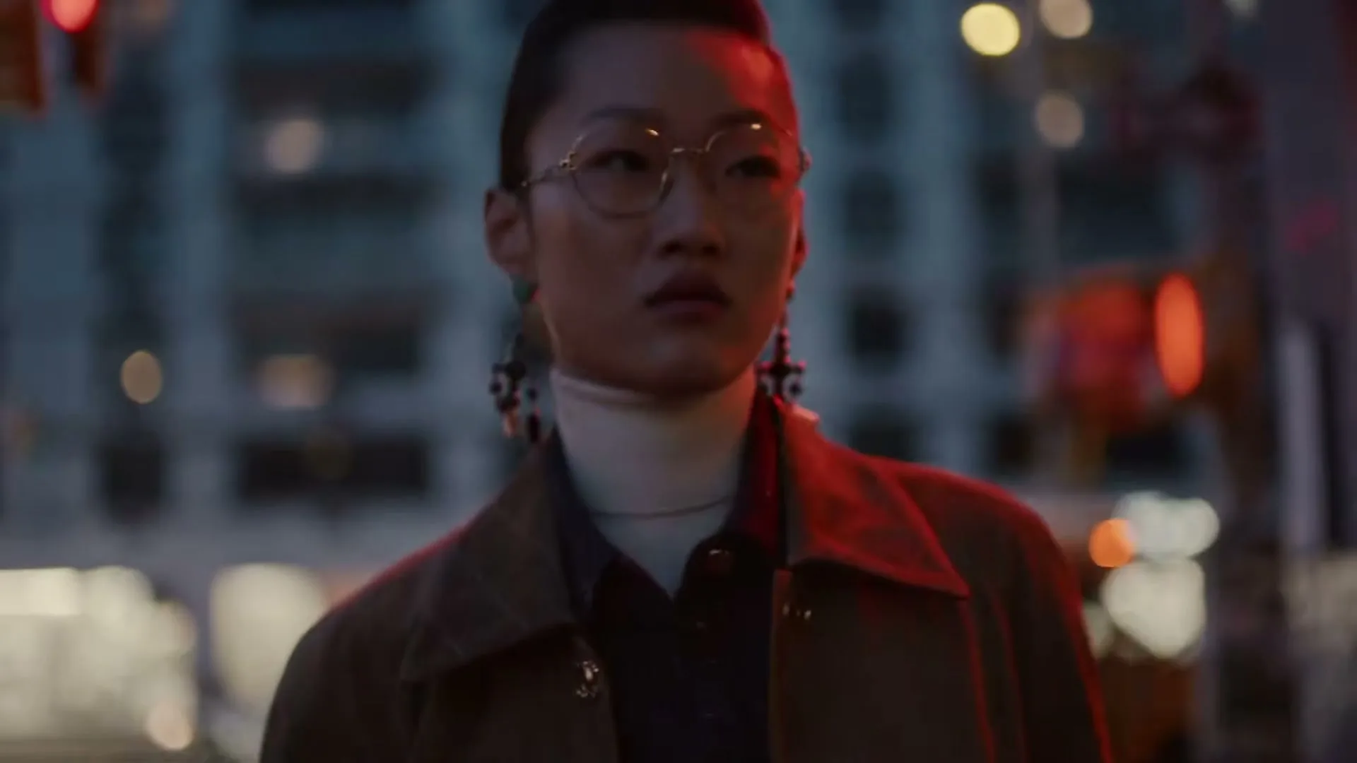 A person with glasses and unique earrings walks towards the camera at night, illuminated by red light from the side, with city lights blurred in the background.
