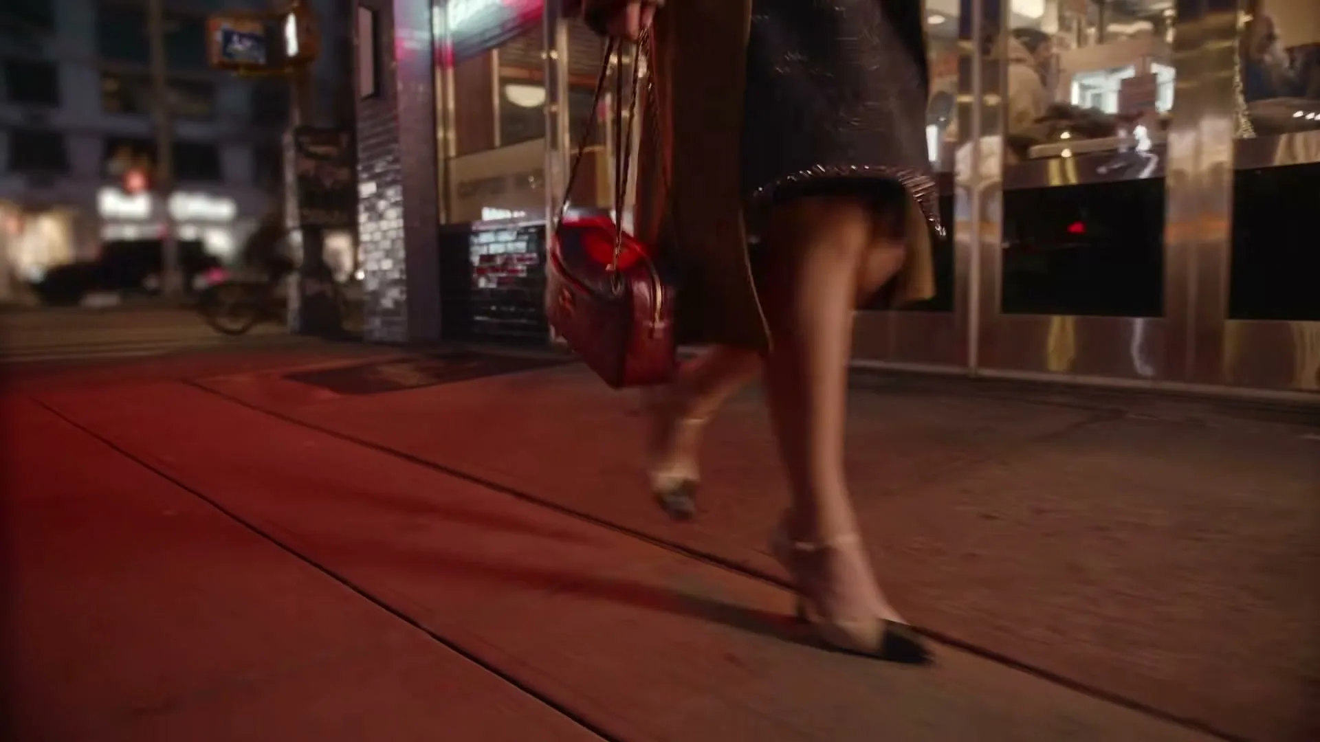 A woman in a black dress and heels walks on a city sidewalk at night, carrying a red handbag.