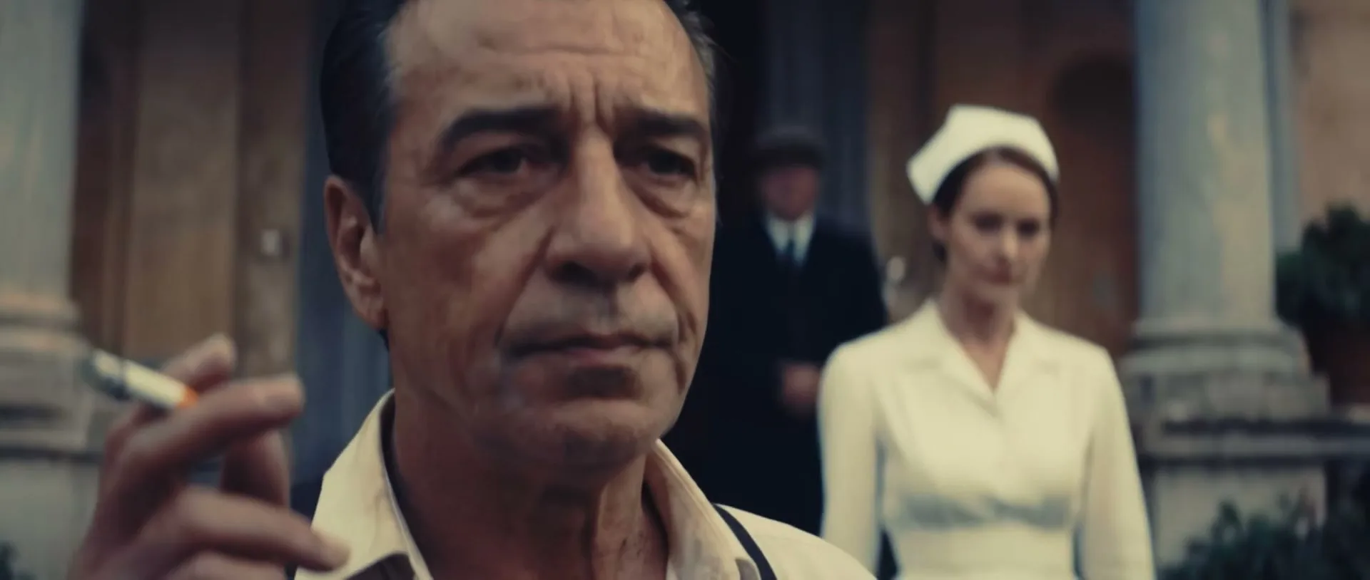 A man in his 50s smokes a cigarette, looking serious, as a woman in a nurse's uniform approaches him outdoors.
