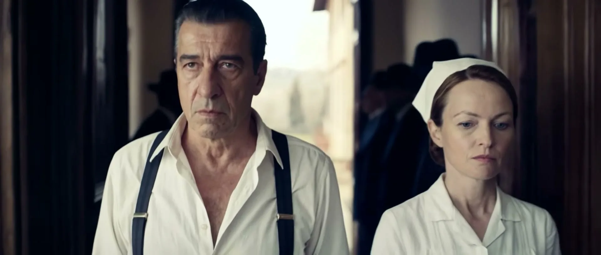 A serious-looking man in a white shirt and suspenders stands next to a female nurse in a white uniform, both looking forward with a determined expression.