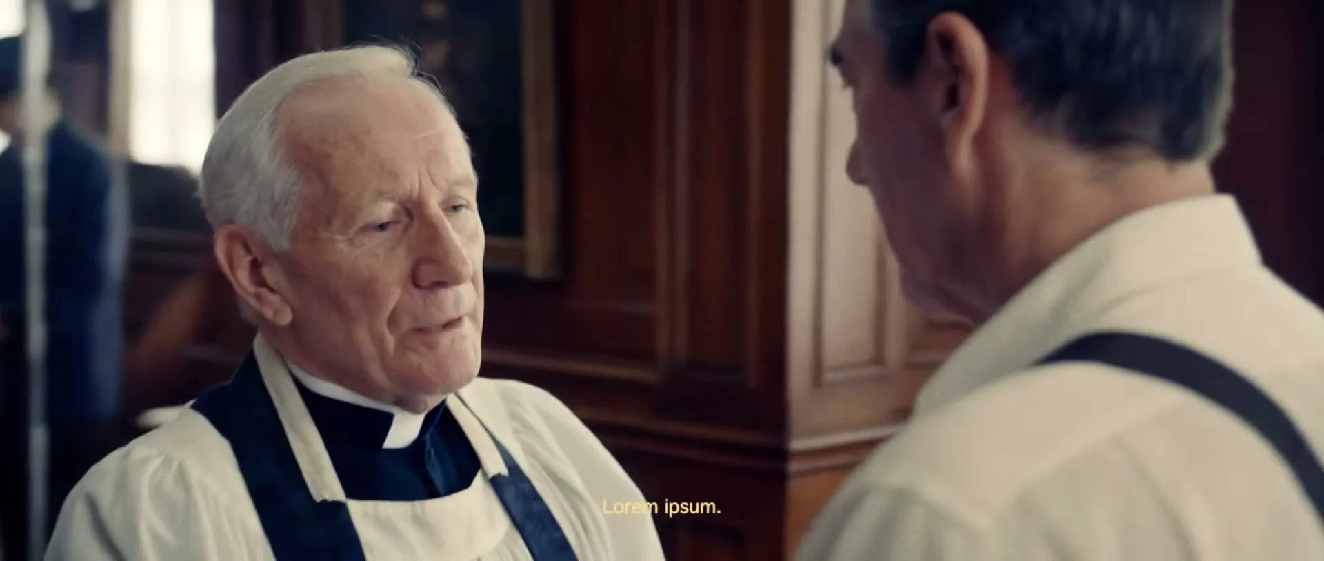 A priest in a white robe and black stole kisses the hand of a man in a white shirt and suspenders, in a room with other people in the background.