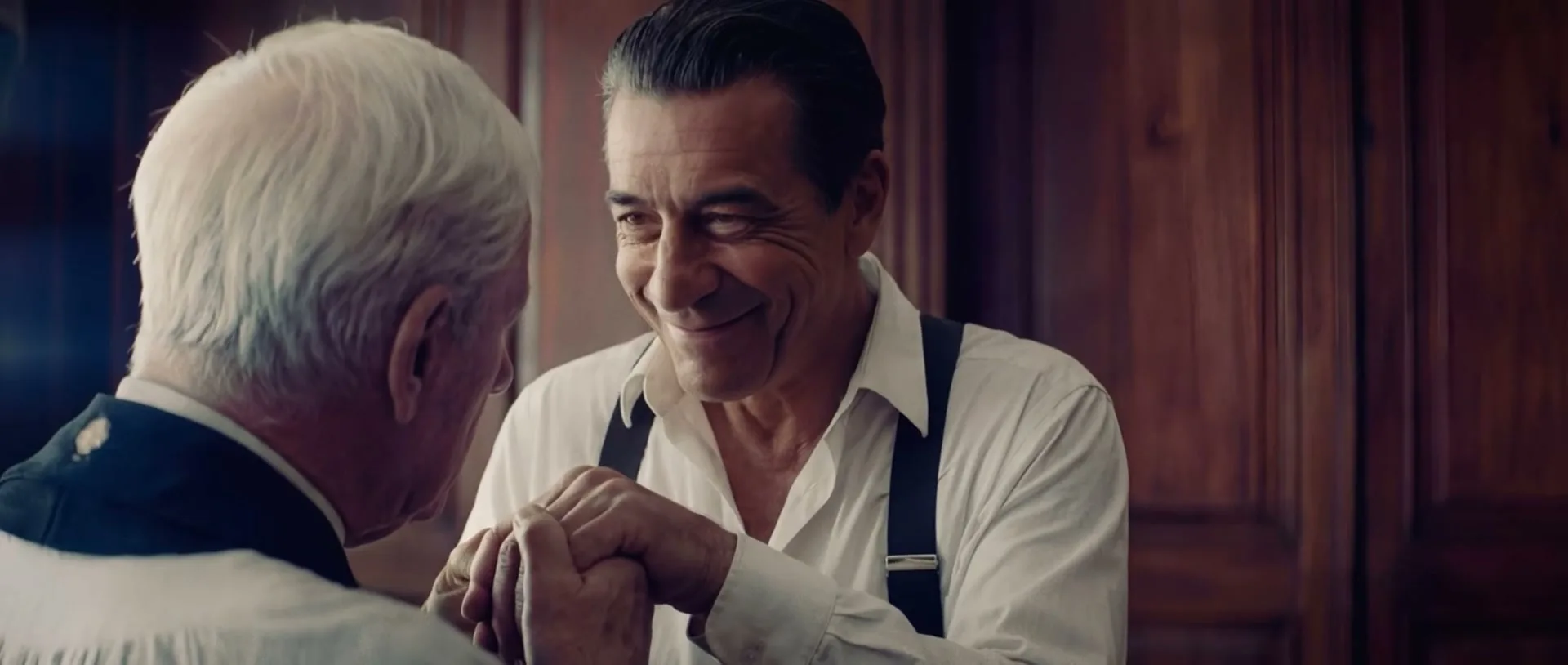 A man with dark hair and a white shirt with suspenders holds the hands of an older man with white hair, smiling warmly in a room with wood paneling.