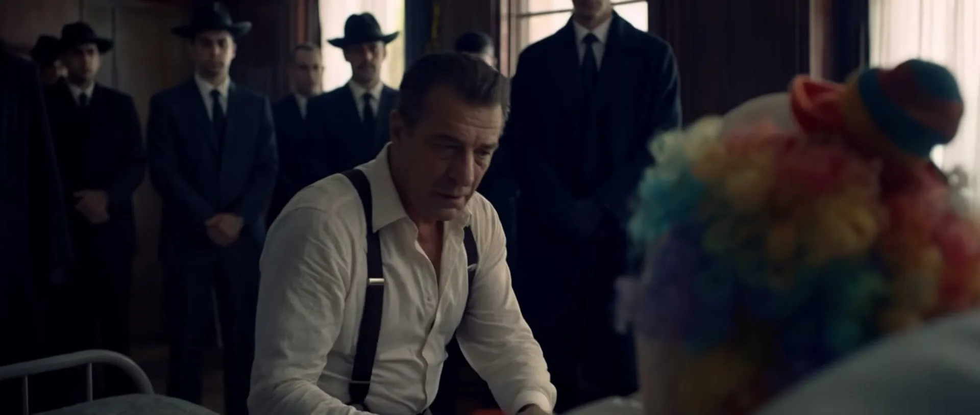 A somber man in a white shirt and suspenders leans over a person in a clown wig, surrounded by men in suits and hats in a dimly lit room.