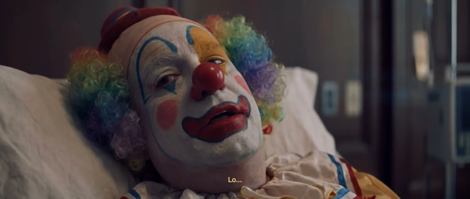 A clown, wearing a colorful wig and makeup, lies in a hospital bed, looking unwell and struggling to speak.