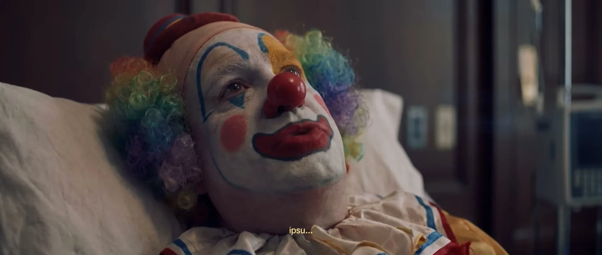 A clown in full makeup and costume lies in a hospital bed, looking weak and unwell, as if on their deathbed.