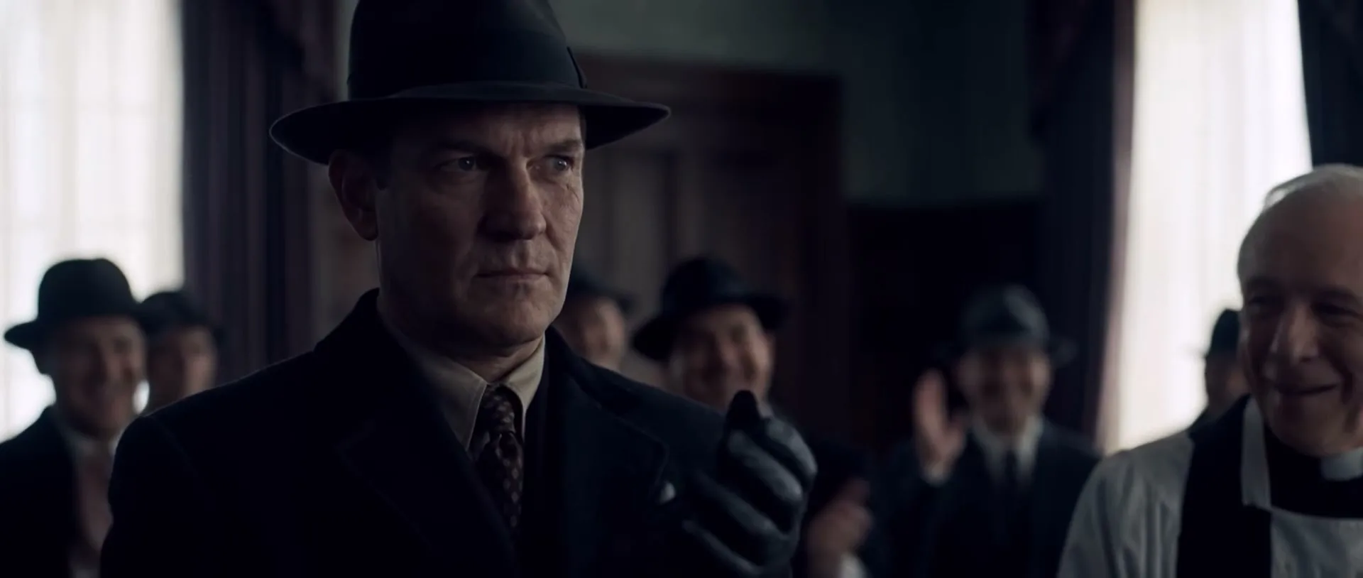 A man in a fedora and suit looks at the camera, then smiles as people in the background applaud him in a dimly lit room.