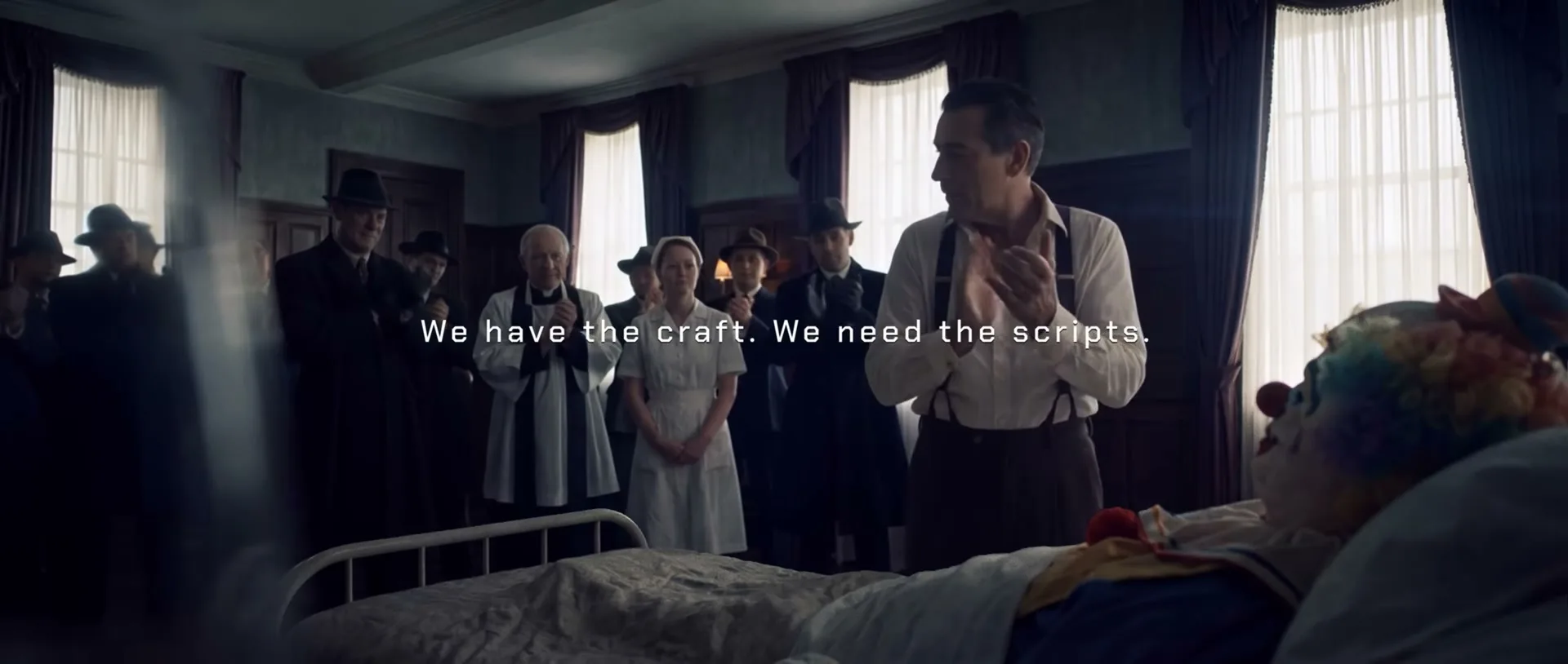 A man in suspenders claps next to a clown in a hospital bed, surrounded by people in a dimly lit room.