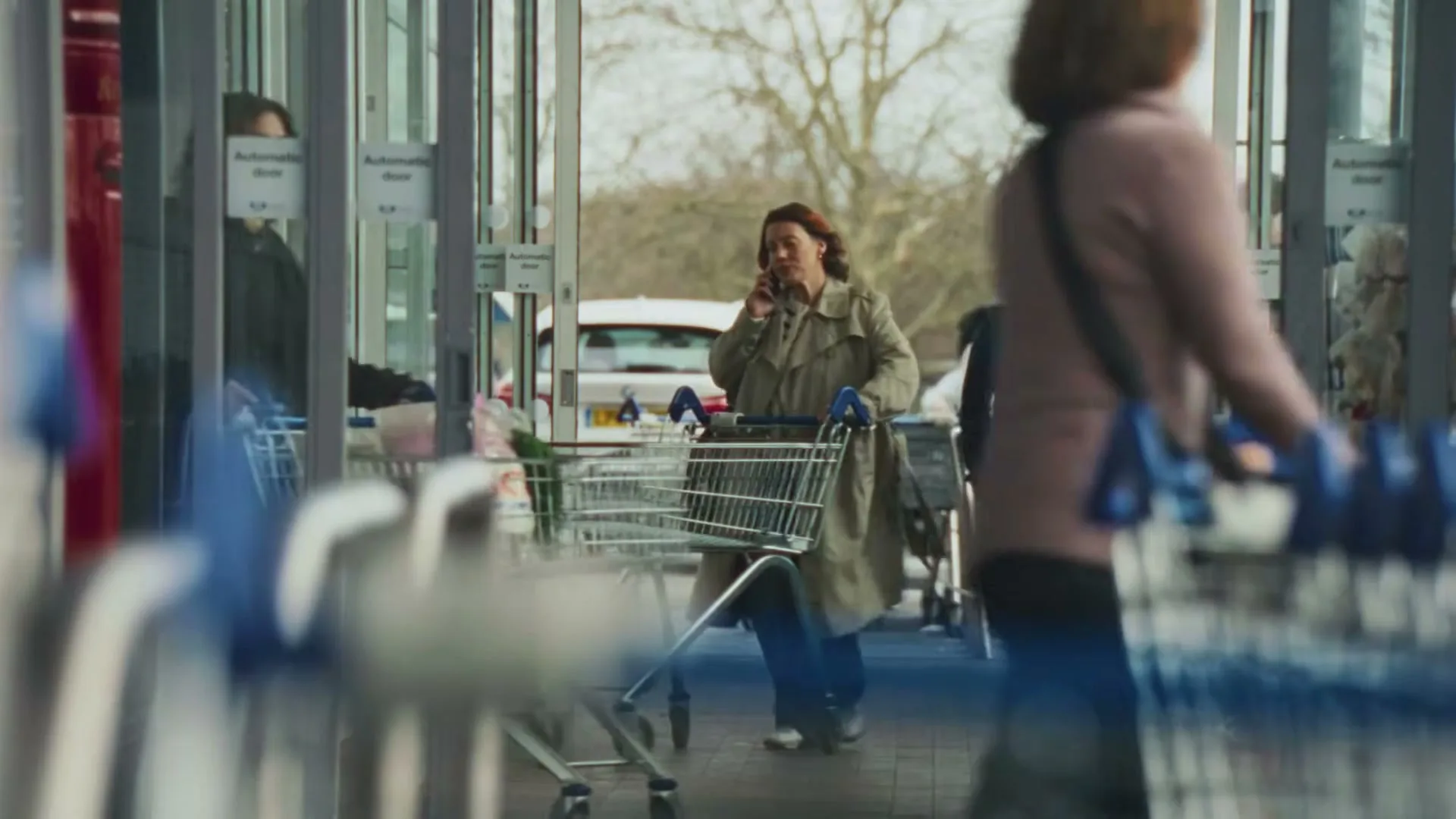 A woman in a trench coat talks on her phone while pushing a shopping cart with a young boy next to her, entering a supermarket.