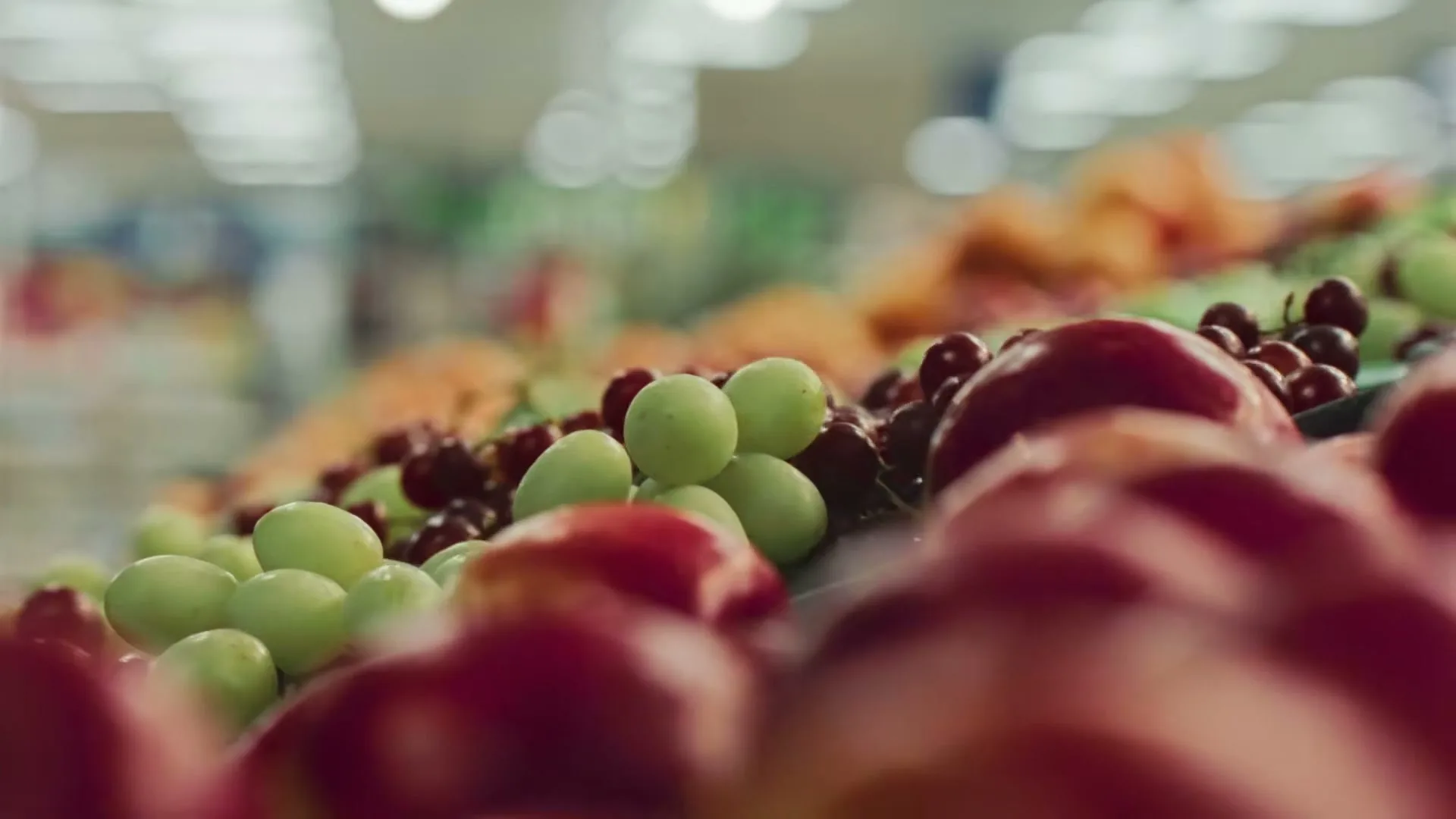 A macro shot of fresh green grapes and red apples on display in a supermarket produce section, with other fruits blurred in the background.