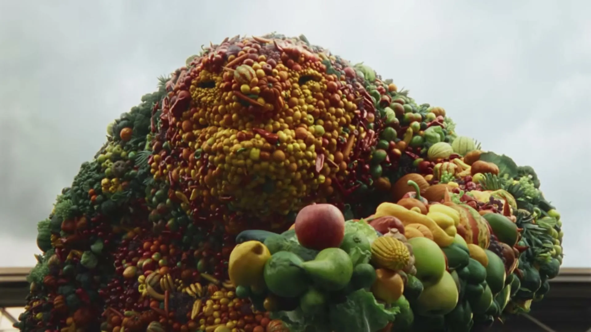 A giant creature made of fruits and vegetables smiles, holding a large pile of produce in its arms, against a cloudy sky.