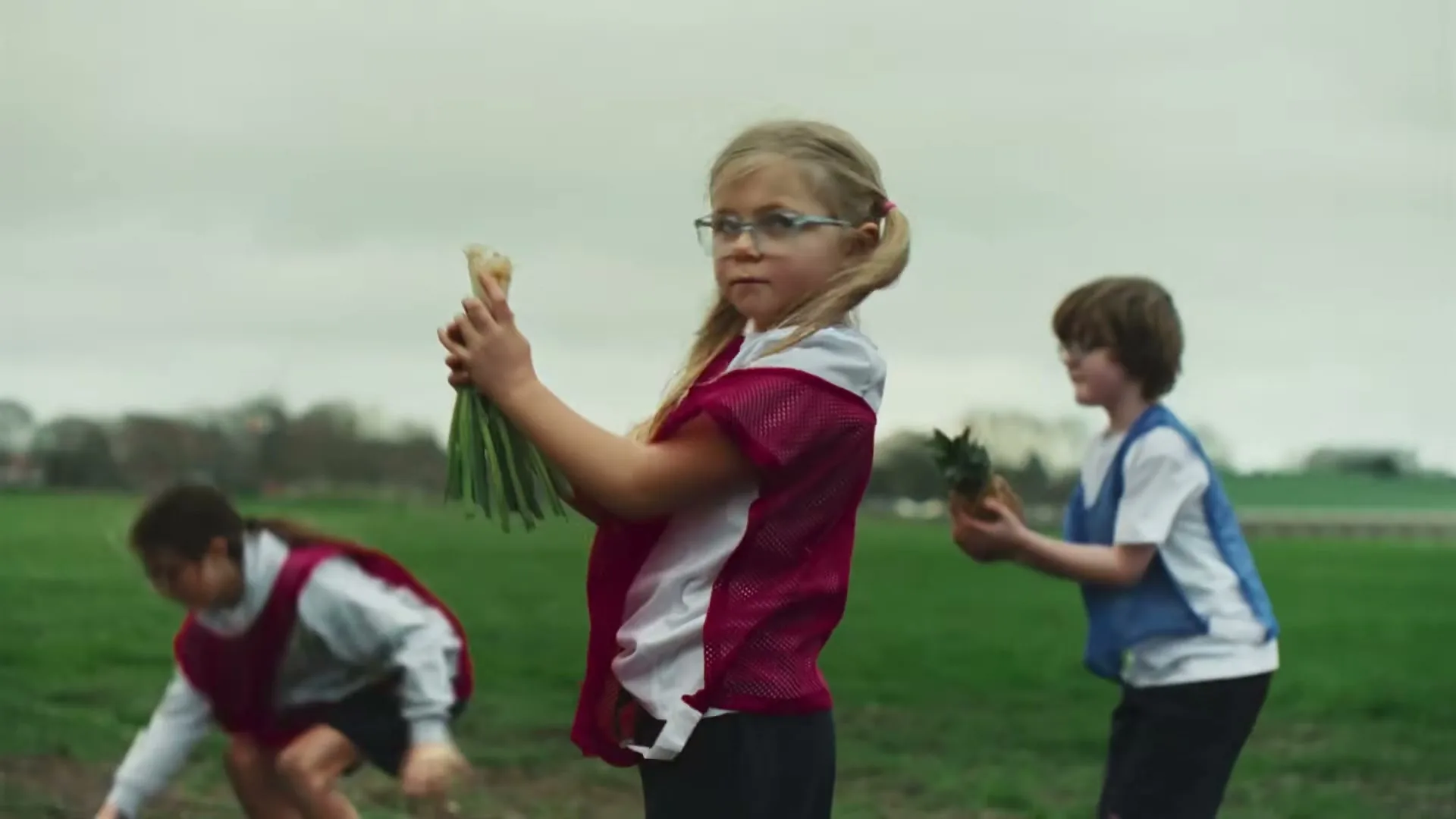 Three children in a field, wearing sports bibs, look up at the sky, then look at the camera holding vegetables.