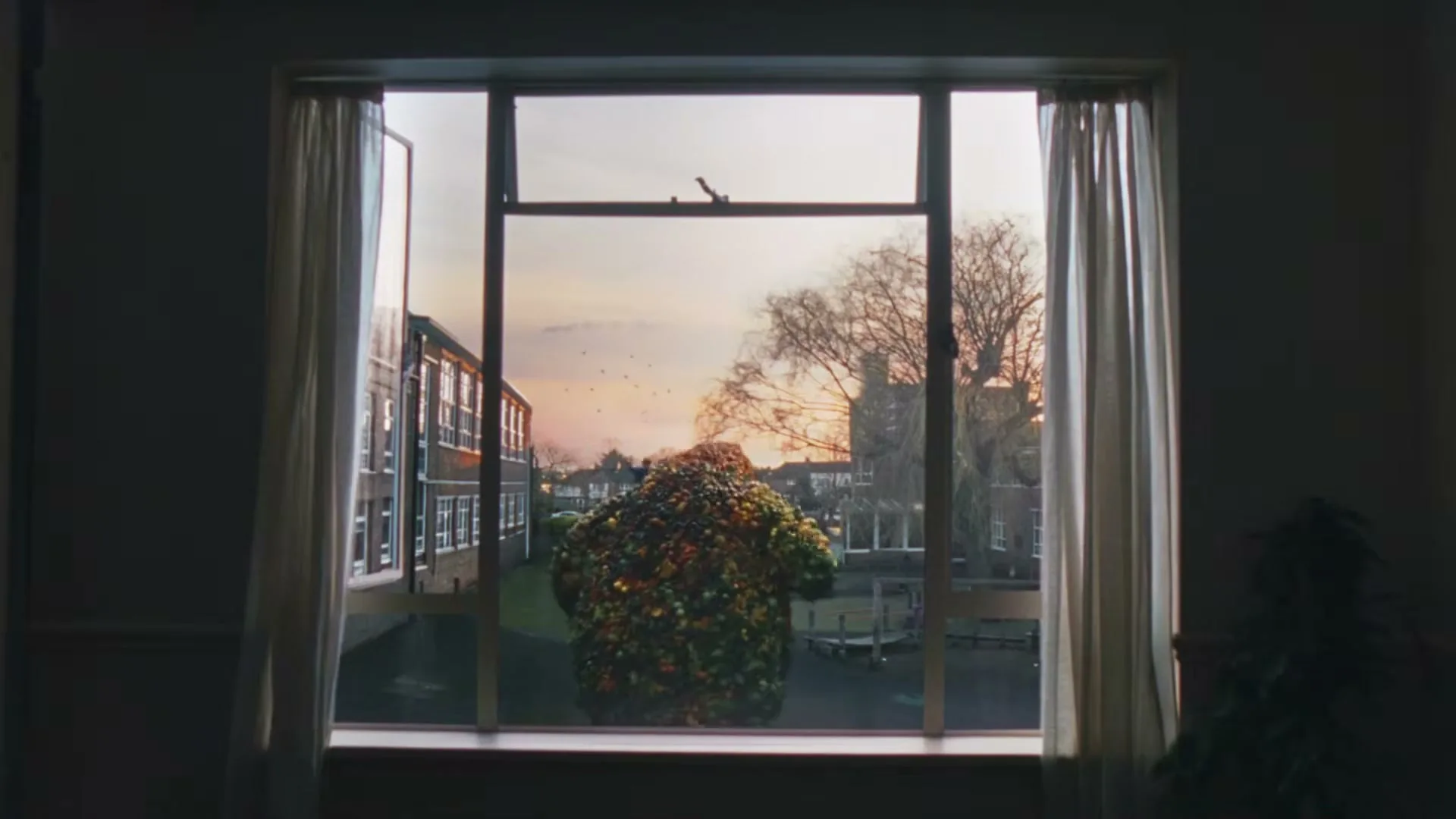 A giant creature made of fruit and vegetables stands outside a window, looking into a building at sunset.