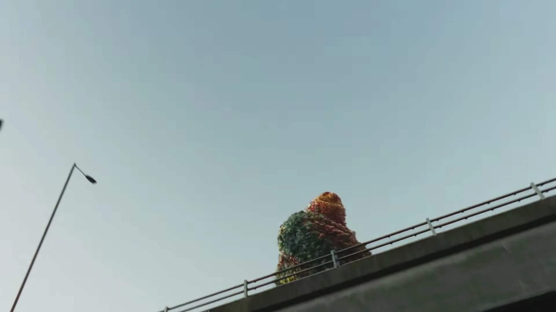 A giant made of fruit walks across a concrete overpass, seen from a low angle against a clear blue sky.