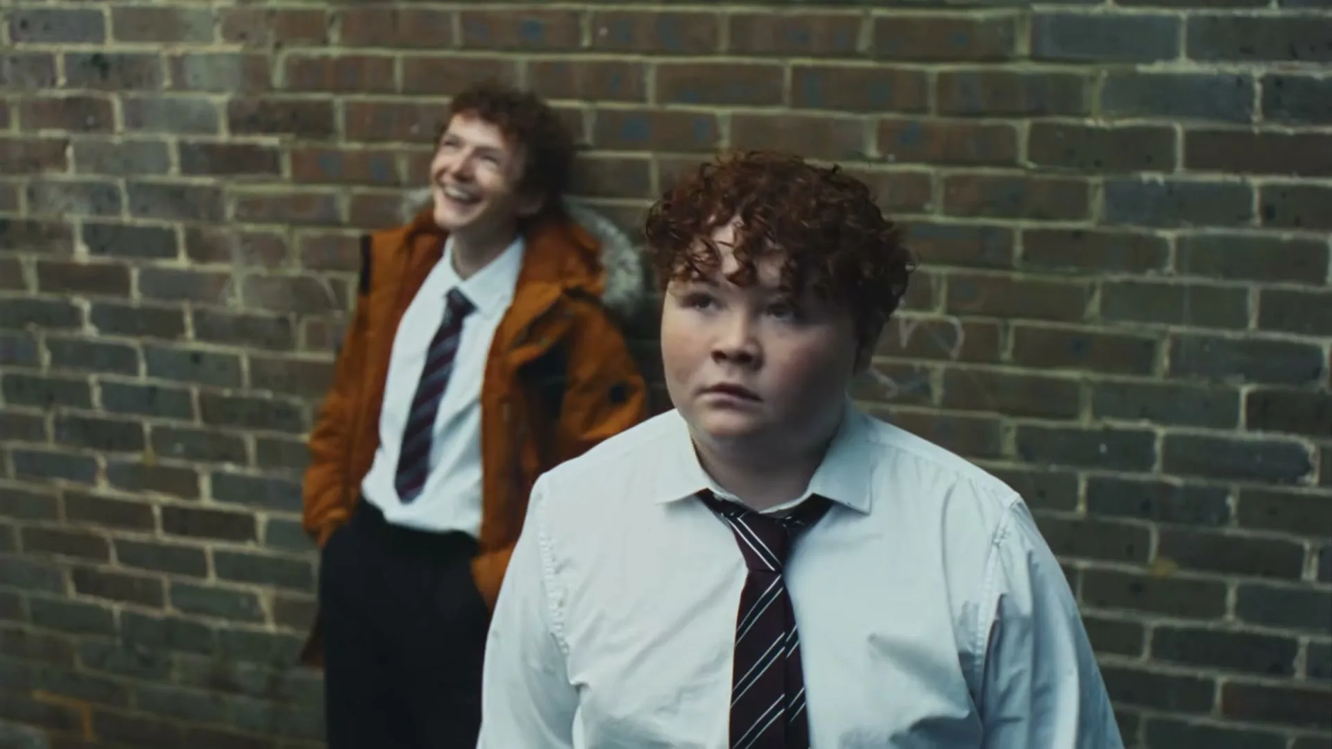 Two male teens in school uniforms stand against a brick wall, one laughing and the other looking away with a neutral expression.