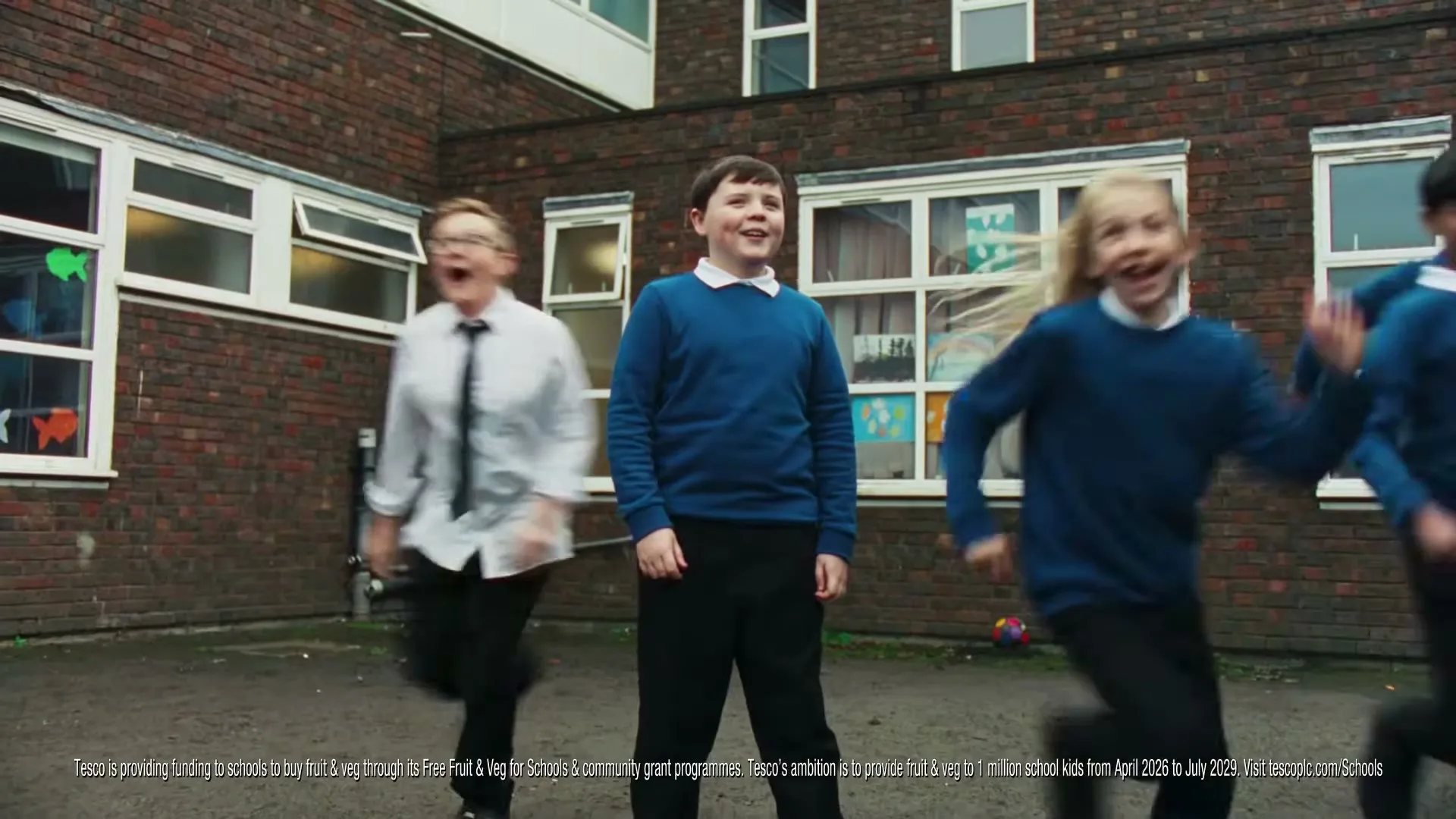 A young boy in a blue jumper stands smiling in a schoolyard as other children run past him, with brick buildings in the background.