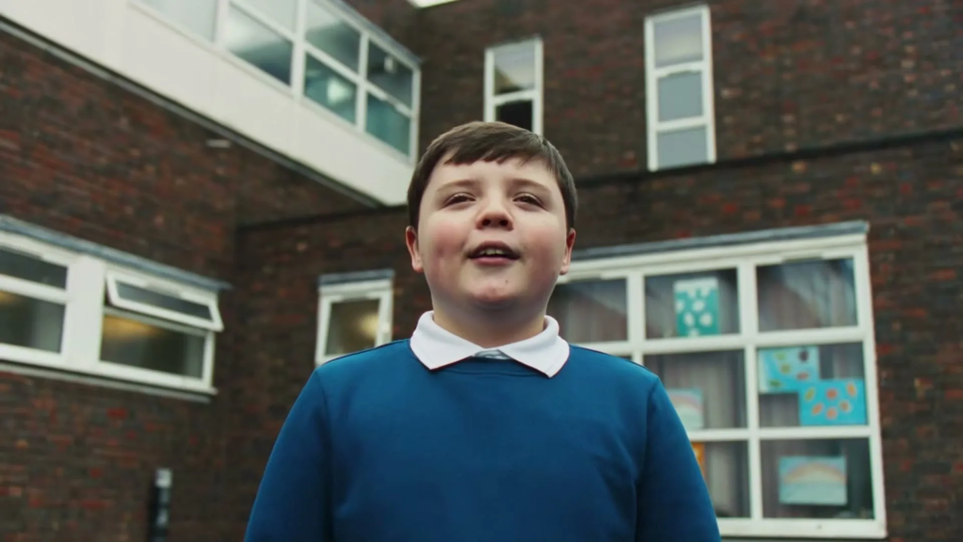 A young boy in a blue school uniform looks up with a curious expression in a schoolyard.