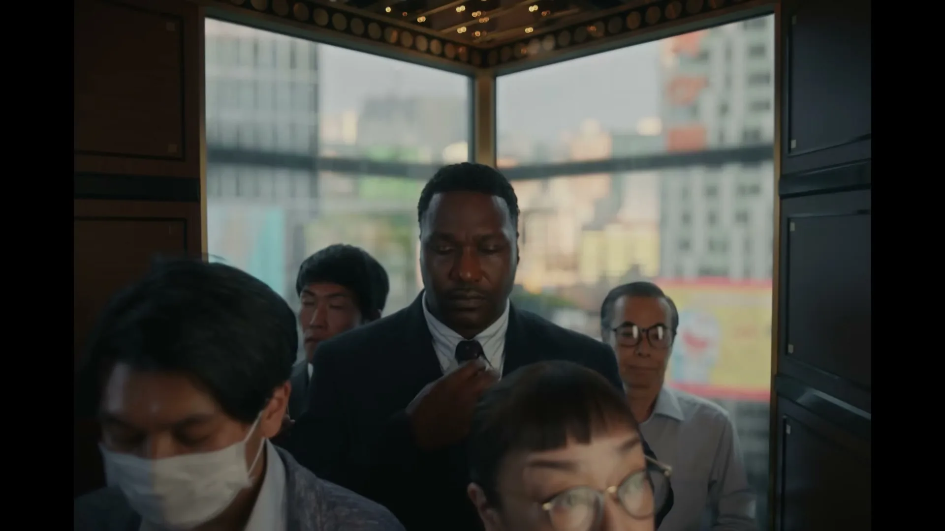 A Black man in a suit adjusts his tie in a crowded glass elevator, surrounded by diverse people, with city buildings visible outside.