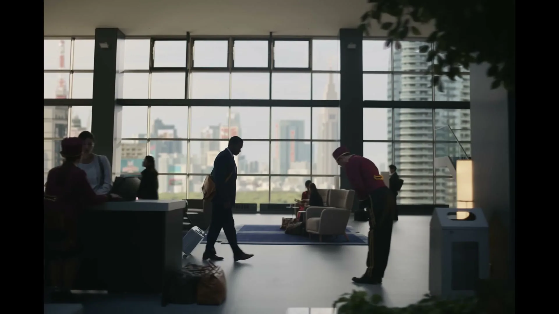 A man in a suit walks through a modern hotel lobby with a cityscape visible through large windows, as a bellhop bows.