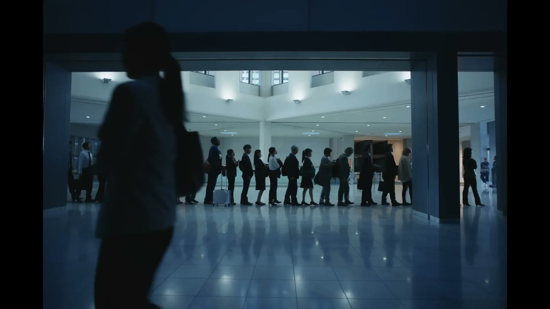 A silhouetted woman walks past a long line of people standing still in a brightly lit, modern interior space, possibly an airport or convention center.