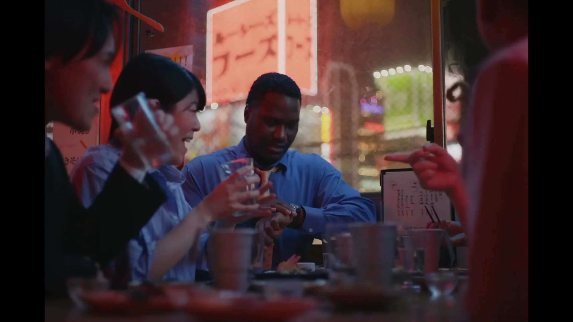A Black man in a blue shirt and orange tie checks his watch while sitting at a table with three Asian people in a dimly lit Japanese restaurant at night.