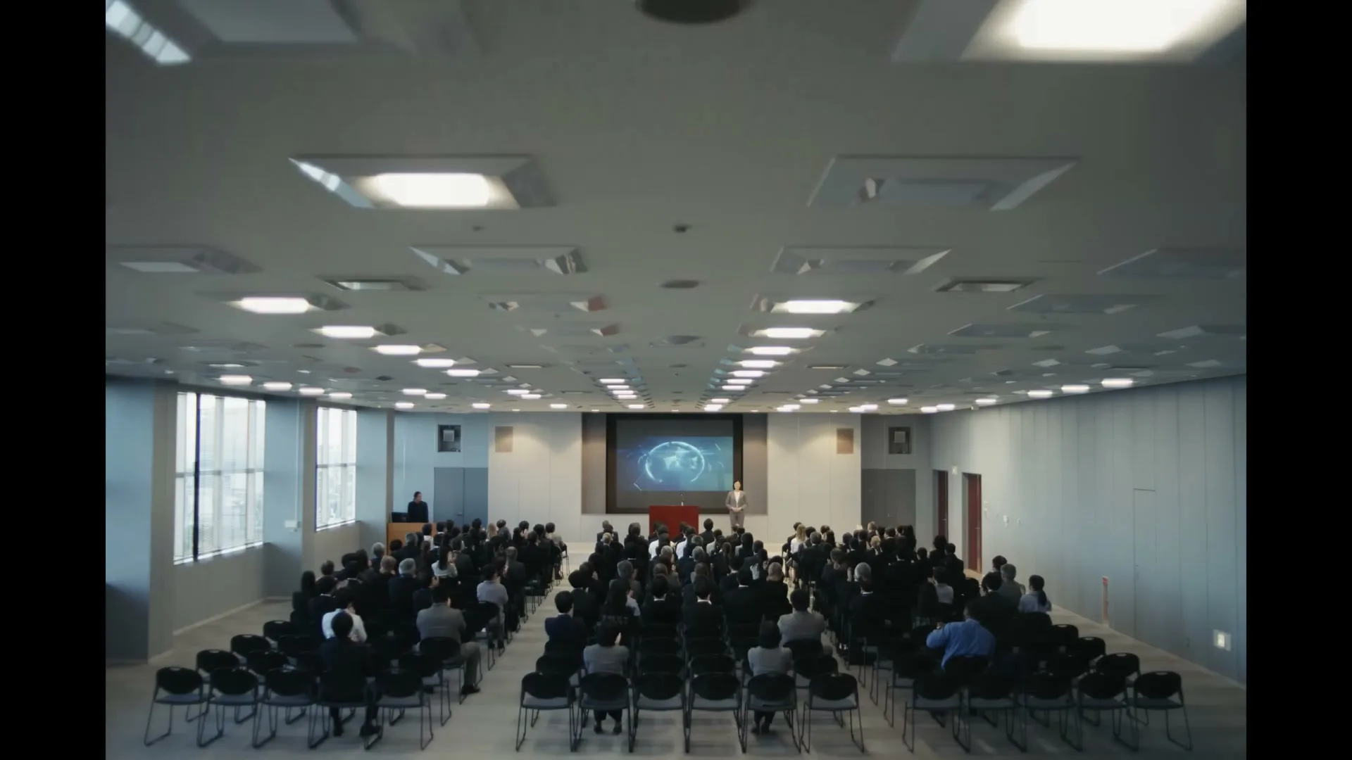 A wide shot of a large audience of people in business attire sitting in a conference room, watching a presentation on a large screen.