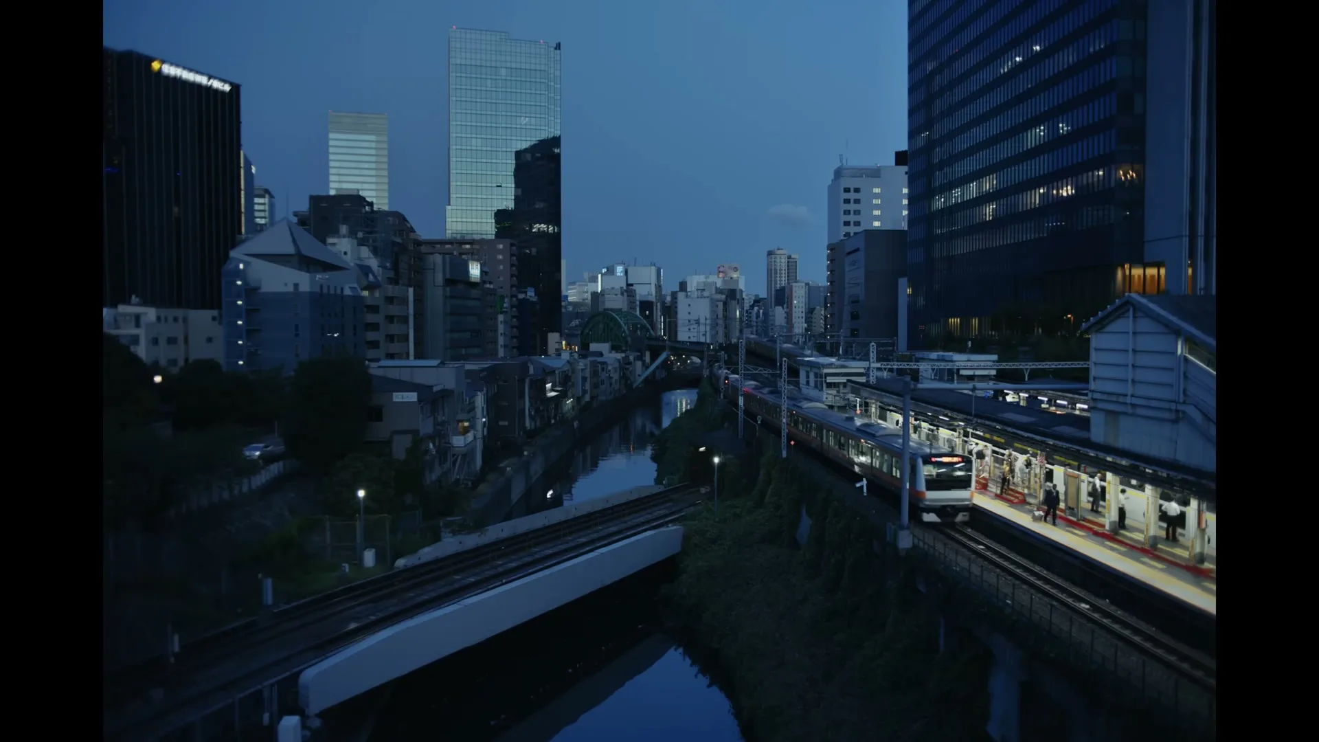 An establishing shot of a Japanese city at dusk, with a train pulling into a station next to a river and tall buildings.
