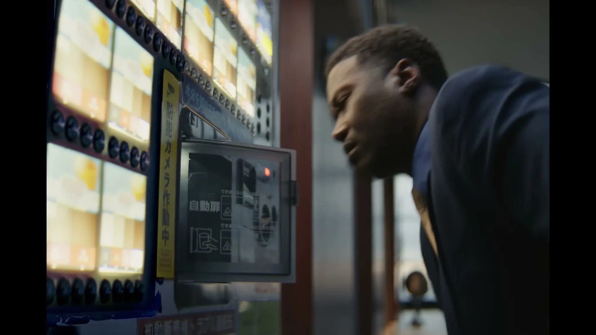 A man in a suit looks closely at a vending machine, his face illuminated by its screen, in a dimly lit interior.