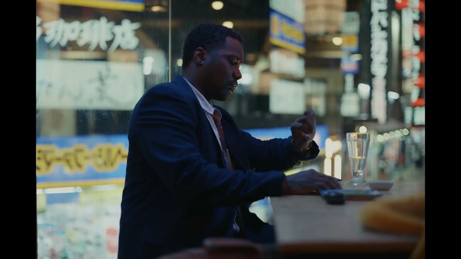 A Black man in a suit sits at a bar counter, looking at his hand, with neon signs visible outside a window at night.