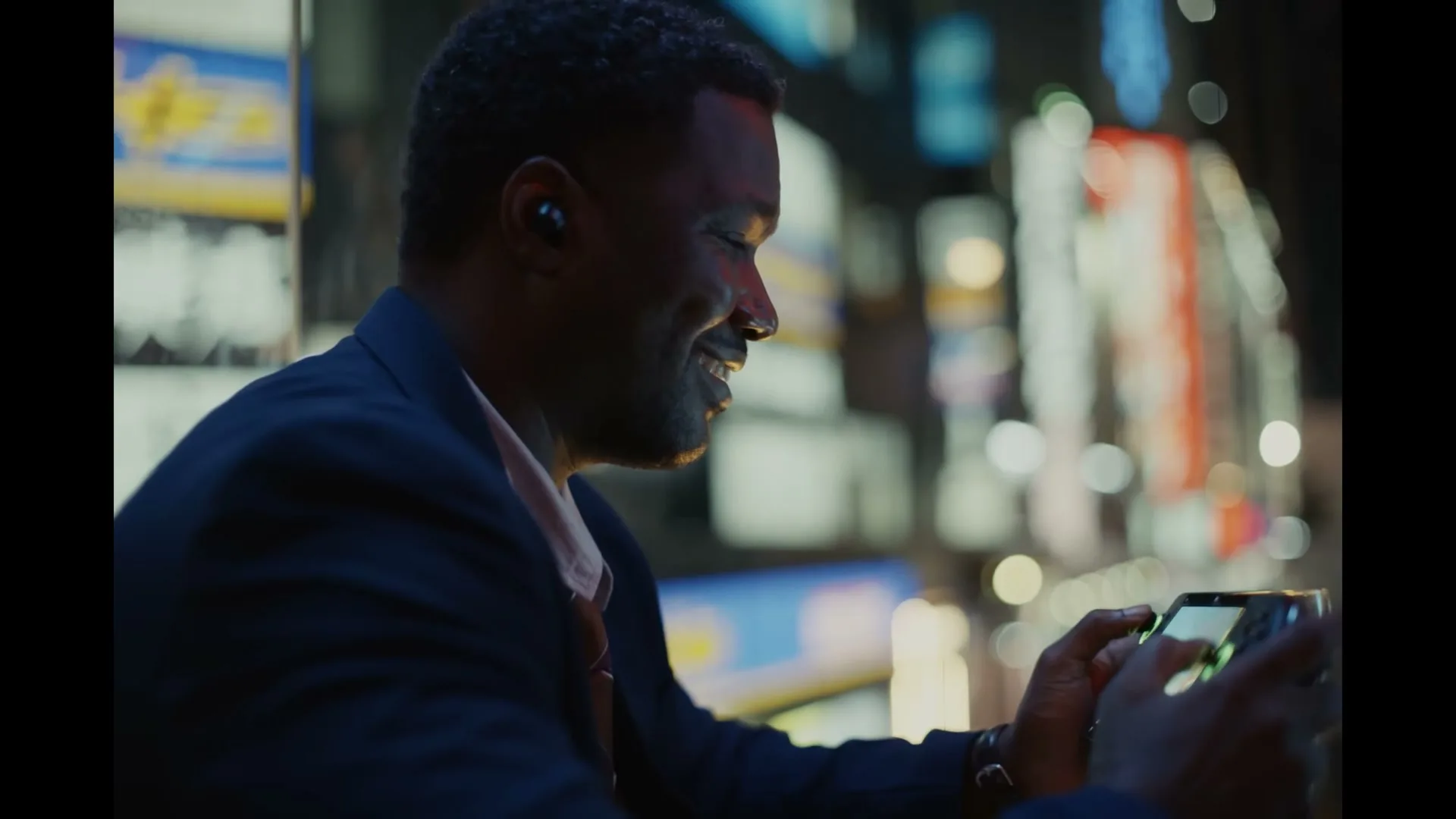 A man in his 30s, wearing a suit and an earbud, smiles while playing a video game on a handheld device at night in a city.