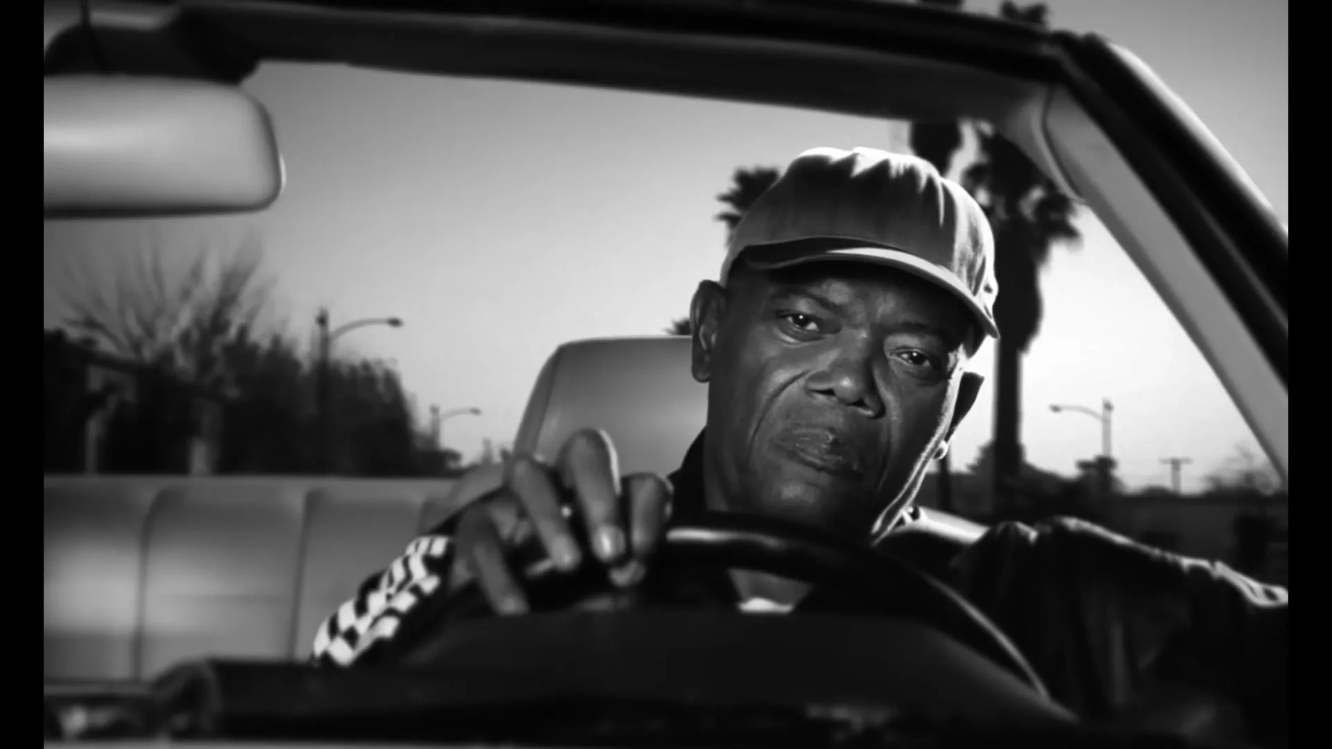 A man in his 60s, wearing a cap and a track jacket, drives a convertible car, looking intently at the road in a black and white shot.