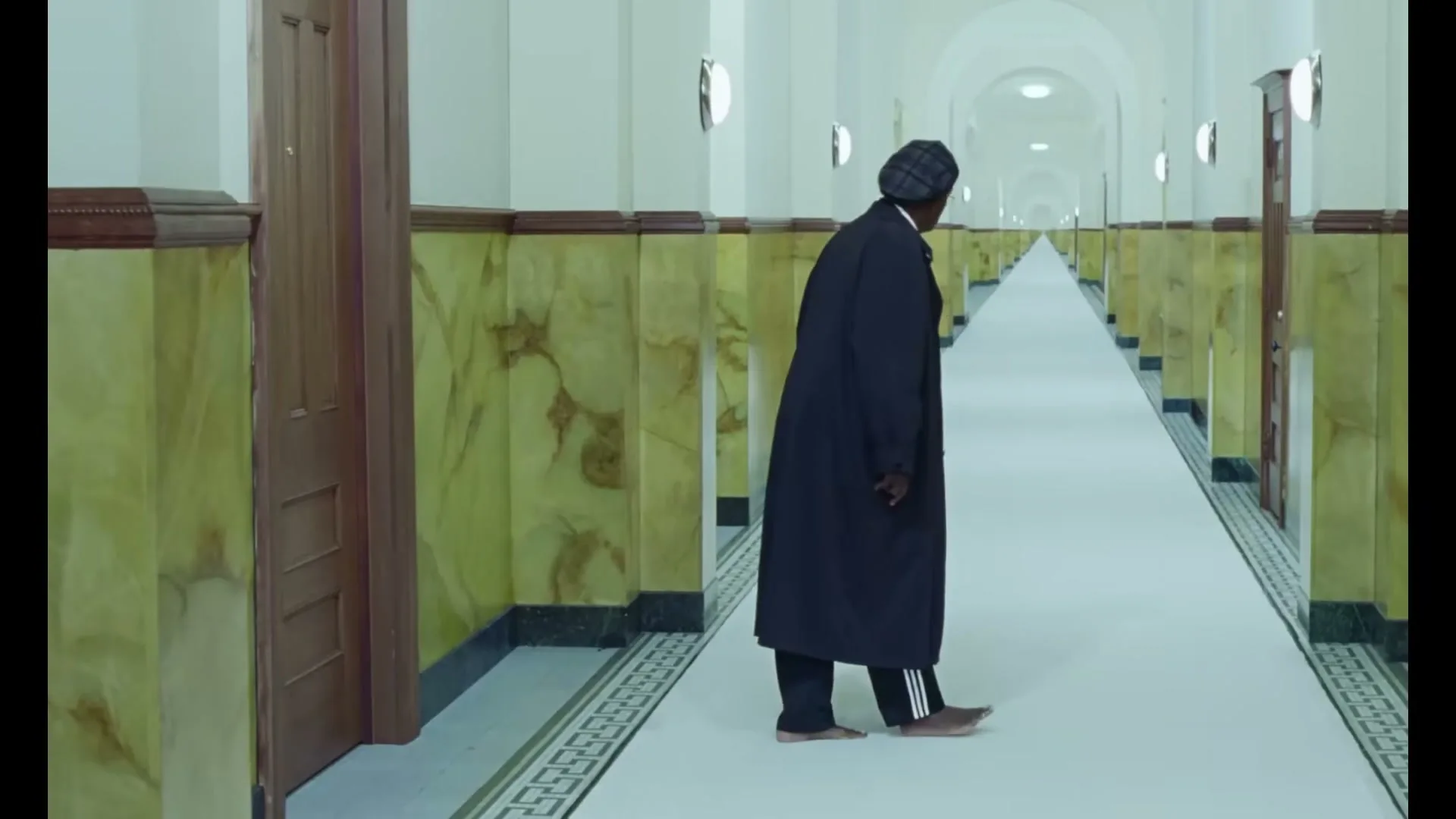 A person in a dark coat and hat walks barefoot down a long, empty hallway with marble walls and many doors.