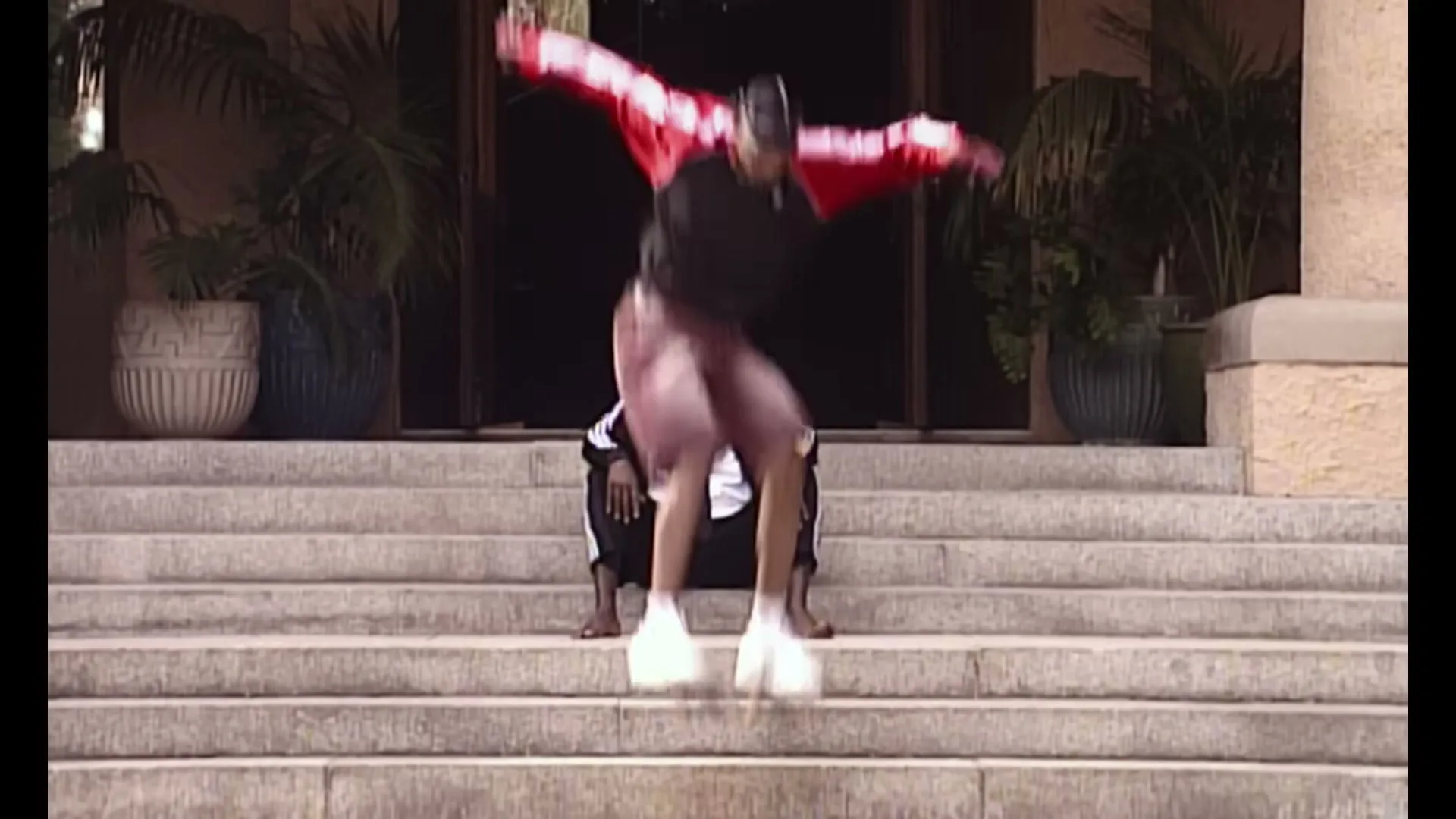 A young man in a red jacket jumps over a seated man in a suit on a skateboard on stone steps outside a building.