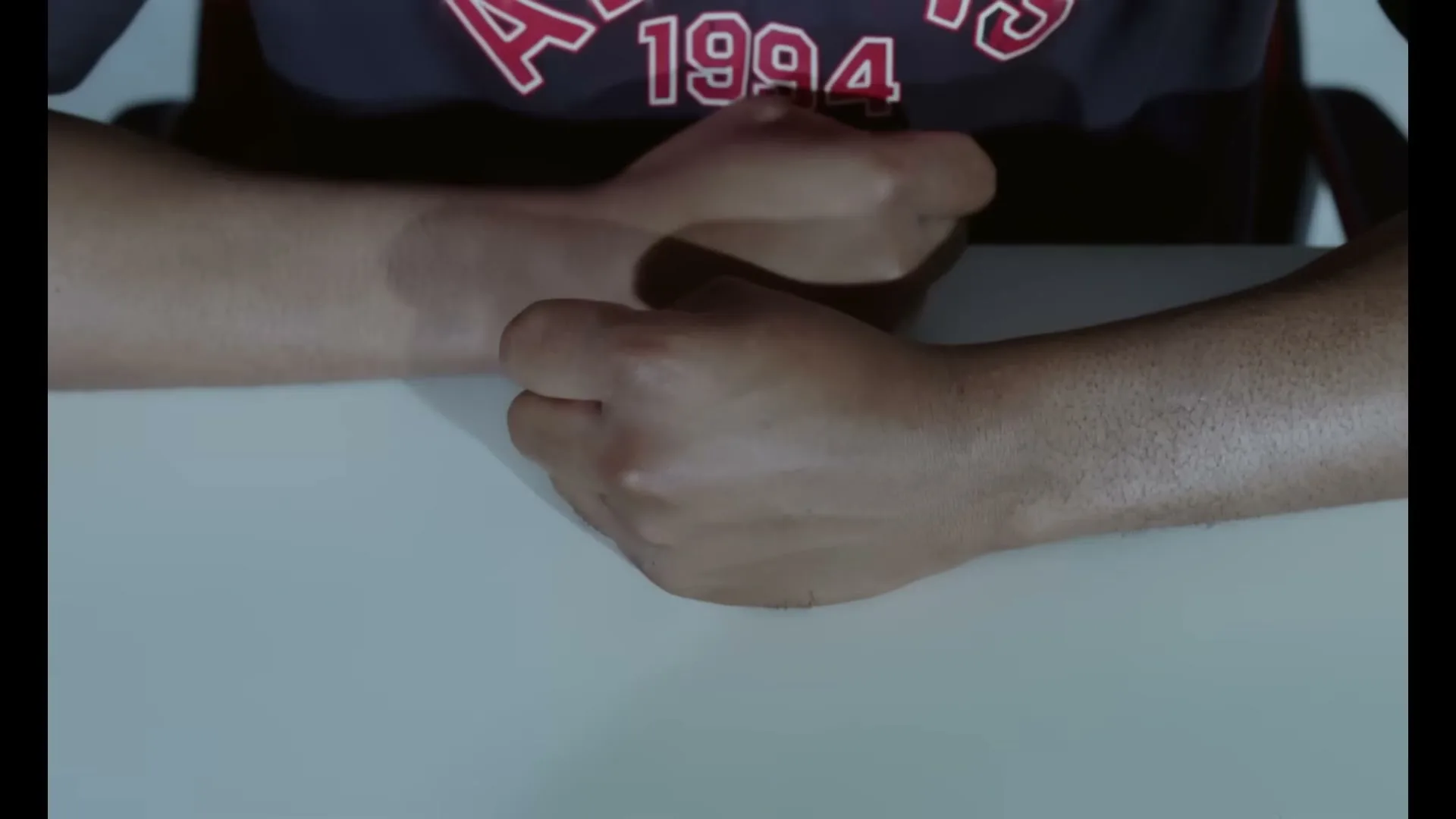 A person with dark skin and a dark t-shirt with red text sits at a white table, clenching and unclenching their fists.